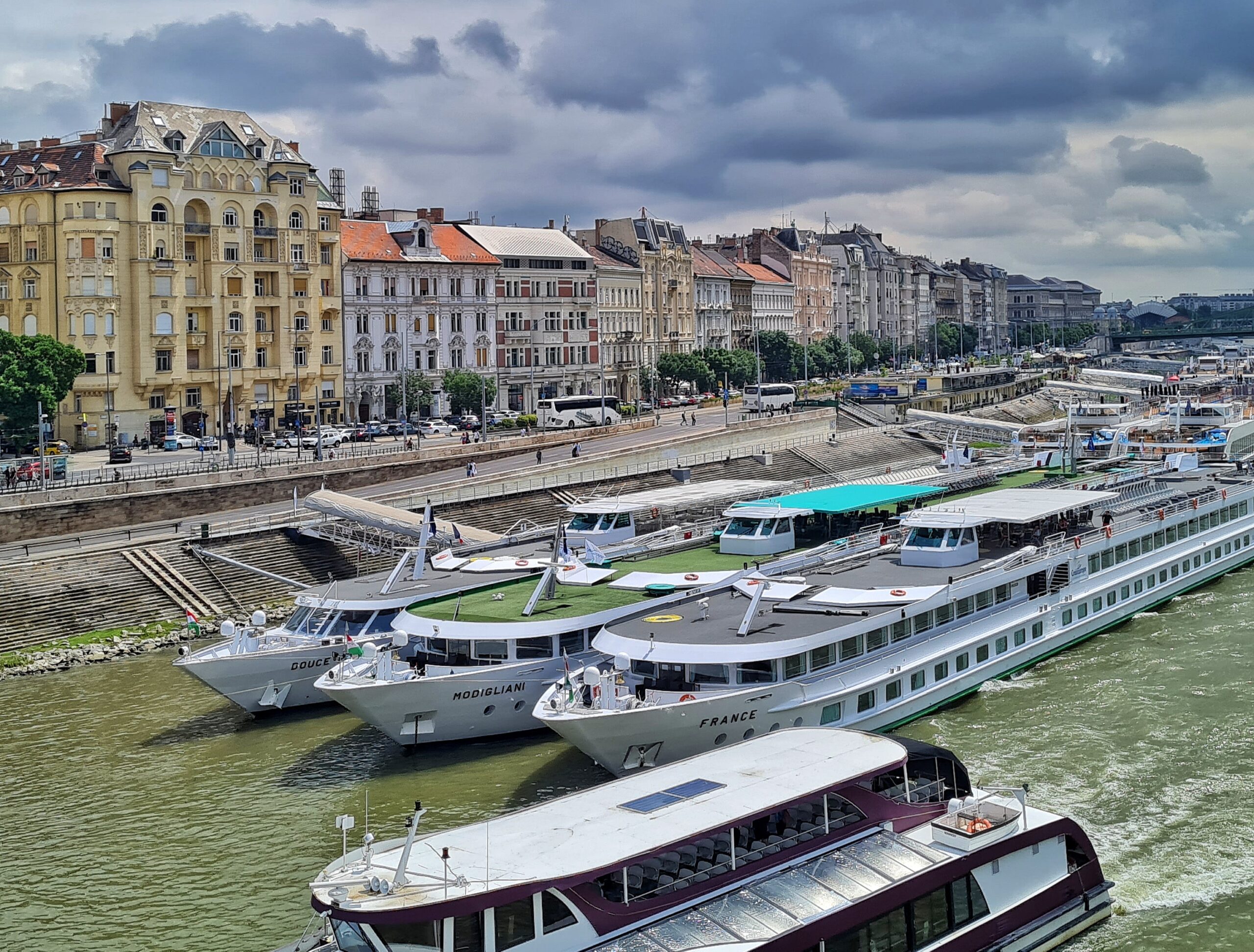 Eine Flußpromenade mit hohen Häusern, Bussen, Pkws und Menschen. Am Ufer liegen nebeneinander mehrere flache Schiffe mit vielen kleinen Fenstern.