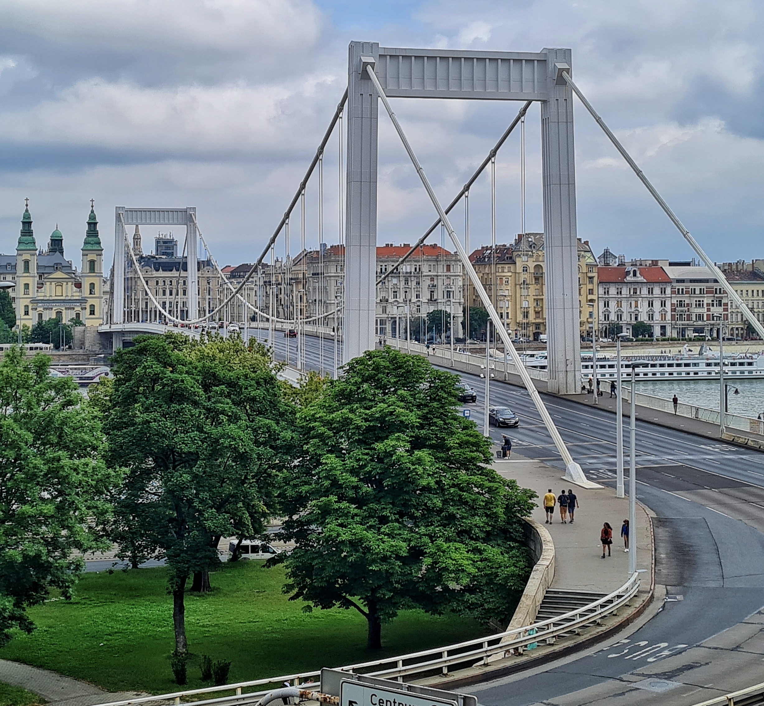 Eine große weiße Brücke. An den beiden Enden hohe viereckige Portale, die durch Seile auf beiden Seiten verbunden sind. Die Seile hängen in der Mitte nach unten, und an den Seilen hängen weitere Seile oder ähnliches nach unten. Auf dem hinteren Ufer viele prächtige Gebäude. Vorne lääuft die Brücke in eine Straße aus, die eine Kurve macht. Hinter der Kurve eine Rasenfläche mit mehreren Bäumen.
