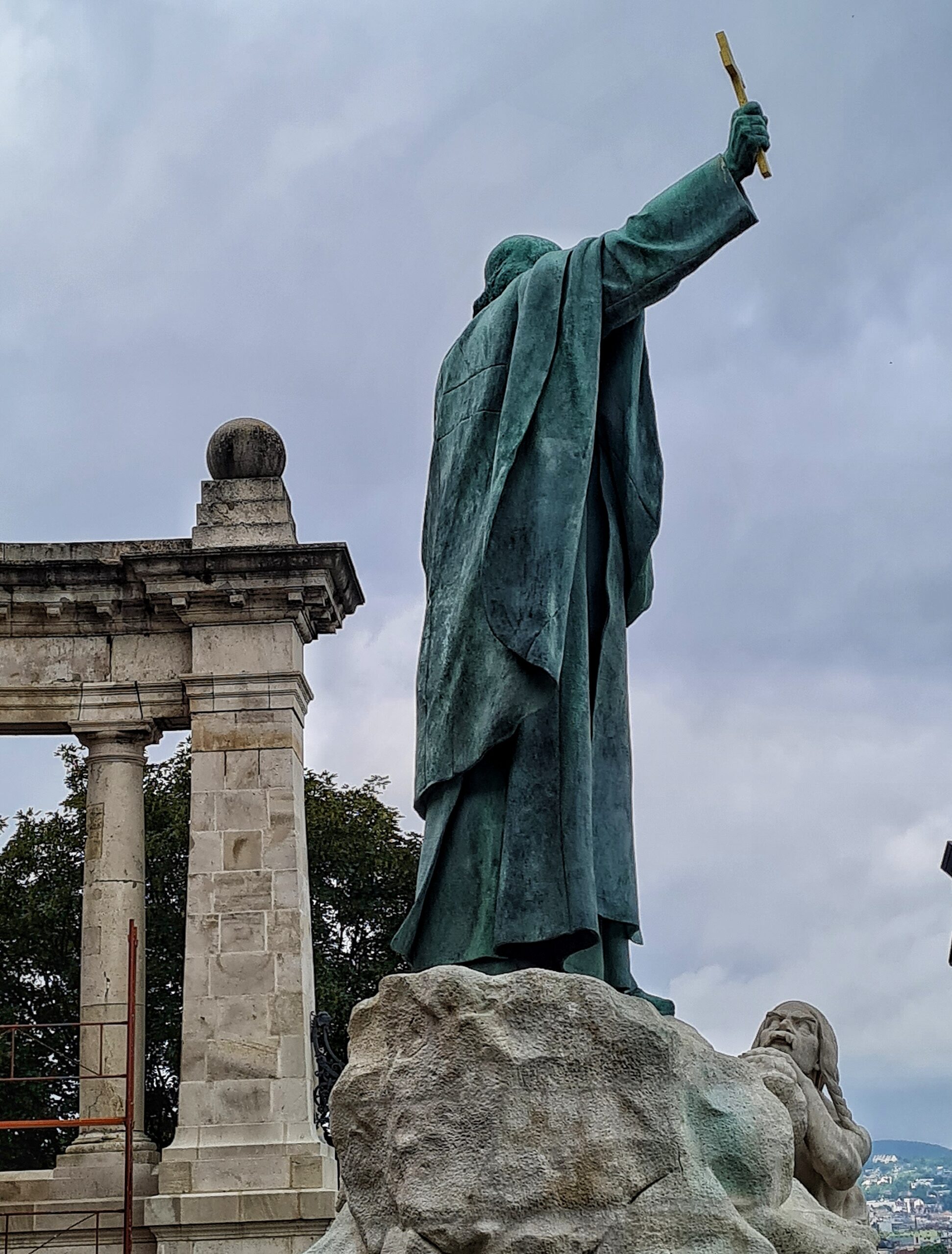 Die Statue aus der Nähe von schräg hinten fotografiert. Sie steht auf einem hellen Stein, an dessen Ende der Kopf und ein Arm eines liegenden Mannes aus Stein zu sehen ist.