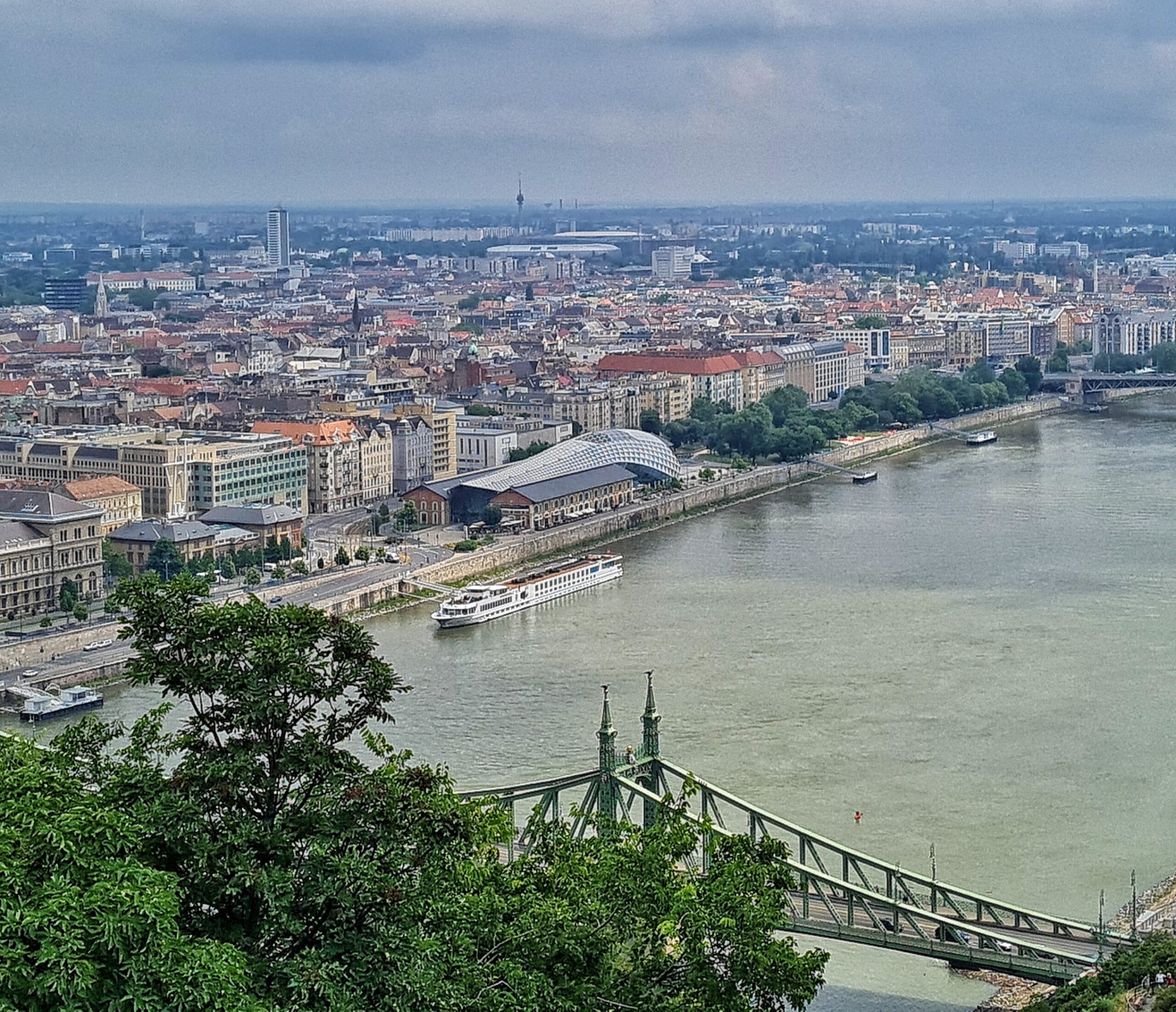 Blick auf die Donai. Im Vordergrund, weitgehend verdeckt durch Bäume eine Brücke mit zwei spitzen Türmen. Auf dewr anderen Uferseite fahren Boote und Schiffe, und es ist die Bebauung von Budapest zu sehen. Auffälig ist ein großes Gebäude am Ufer, das mit einem gebogenen Dach vollständig bedeckt ist.