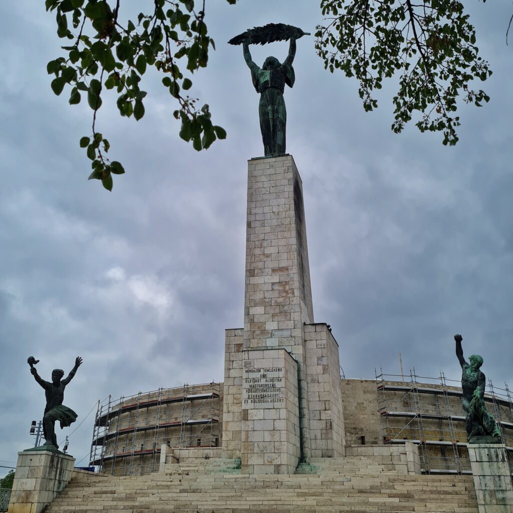 Einige Stufen führen zu einem hohen weißen Turm empor. Rechts und links stehen auf Podesten Bronezstatuen. Links ein Mensch, der beide Arme nach oben hält und in einer Hand eine Fackel trägt. Rechts ein Mann, der einen Arm mit Faust drohend nach oben hält, so als ob er gleich zuschlagen wird. Hinter dem Turm ein flaches rundes Gebäude mit Baugerüsten. Auf dem Turm eine Frauenstatue, die einen großes Blatt quer nach oben hält. Oben links und rechts ragen Zweige mit Blättern ins Foto.