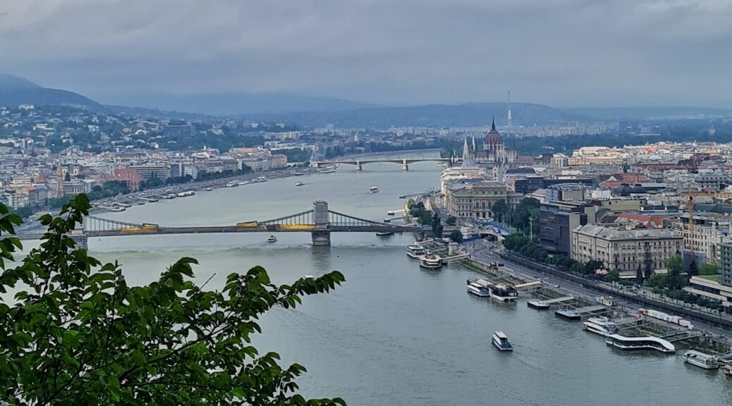 Blick von oben auf einen Fluß, der eine Biegung macht. Im Vordergrund links etwas grünes Gebüsch. Beide Ufer des Flußes sind mit hohen Grbäuden, teilweise mit Türmen,dicht bebaut. Über den Fluß gehen zwei Brücken mit jeweils zwei Brückenpfeilern, eine in der Mitte des Fotos, eine im Hintergrund. Vier kleine Boote fahre auf dem Fluß, an den Ufern liegen weitere Boote und Schiffe.