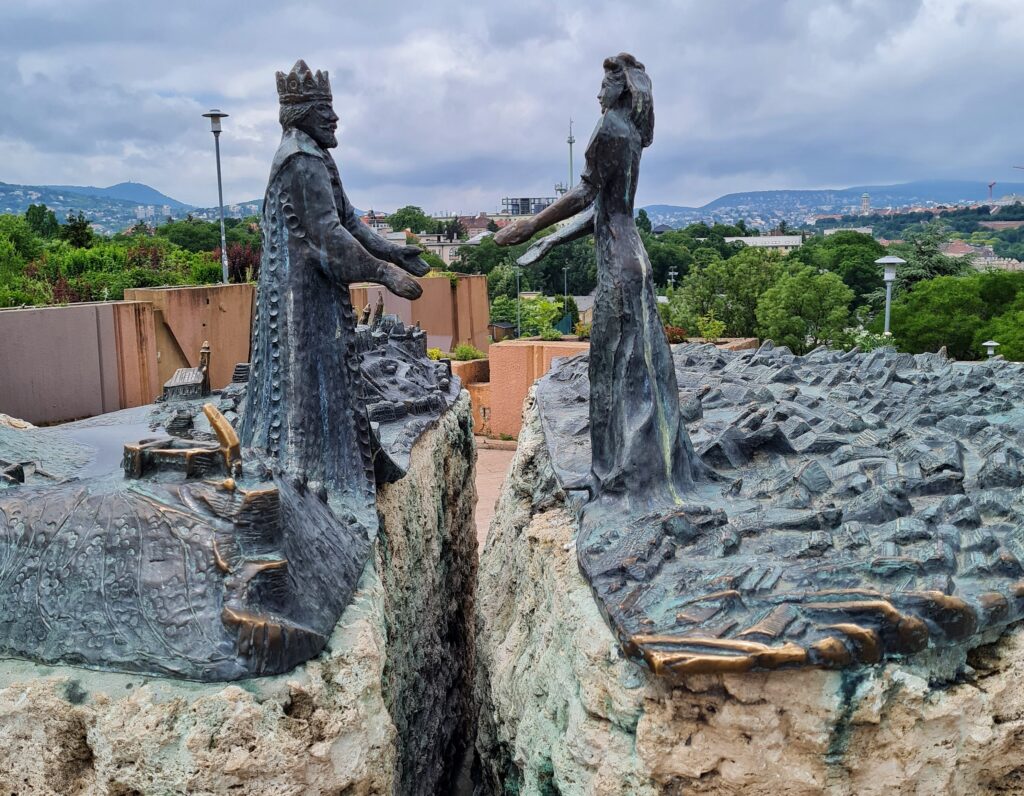 Ein Felsen, in der Mitte gespalten. Darauf eine Metallplatte, auf der Gebäude und Erhebungen aus Metall dargestellt sind. Darauf stehen (links und rechts von der Spalte) zwei Figuren aus Metall, die die Hände zueinander ausstrecken. Links ein König in einem langen Mantel und mit Krone. Rechts eine Frau in einem langen Kleid.