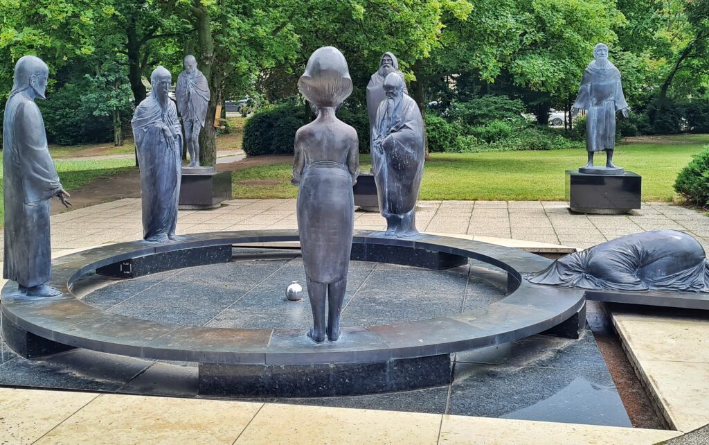 Eine Parkanlage. Darin ein kreisrundes Becken aus Metall. In der Mitte eine Kugel. Um das Becken herum vier stehende Statuen. Eine weitere liegt auf dem Boden und hat ein Tuch vollständig über den Körper gezogen. Im Hintergrund stehen drei weitere Statuen.