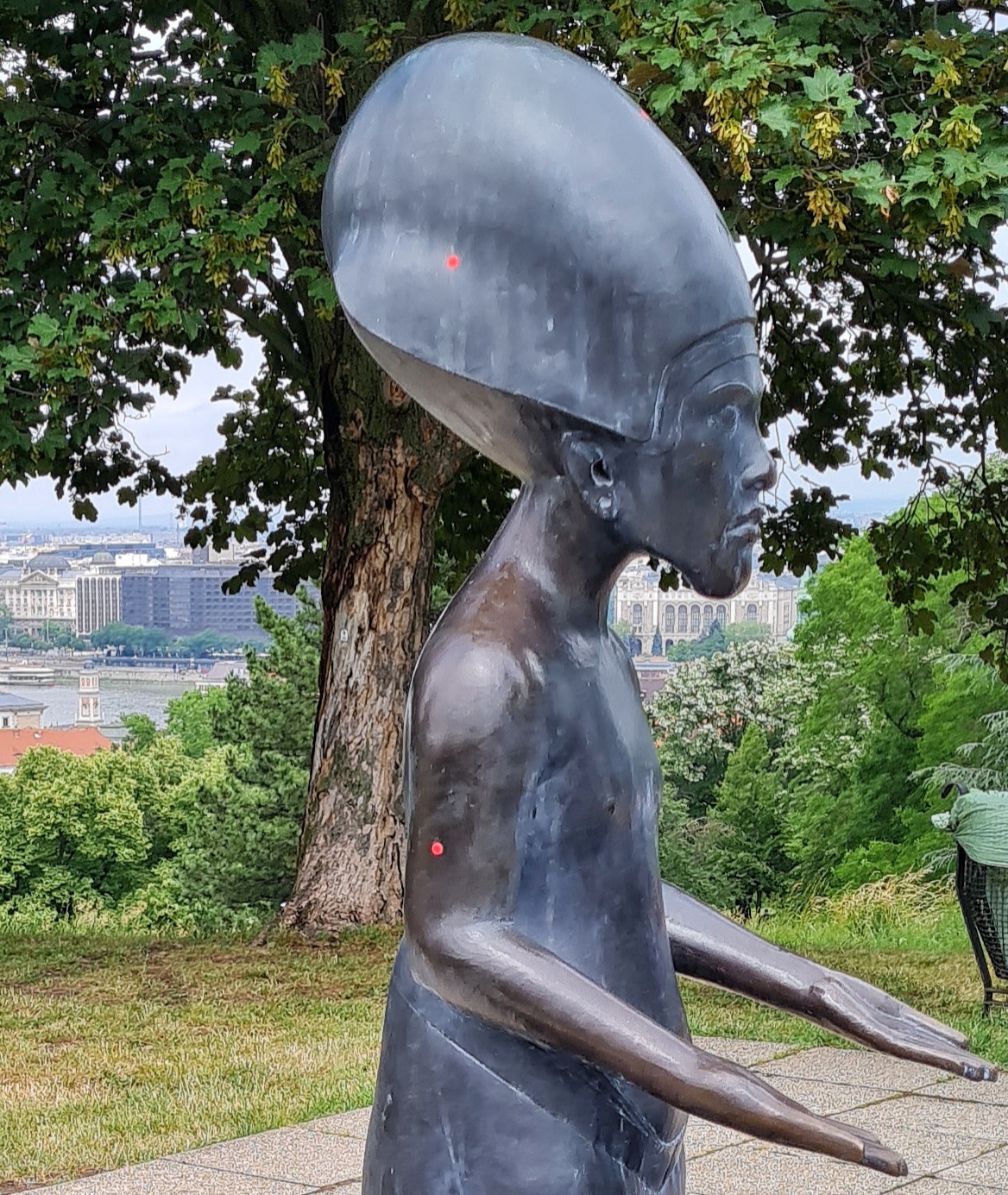 Eine Statue von der Seite.Sie hat eine riesige Kopfbedeckung, so dass sie ein bisschen wie ein Alien aussieht. Sie hat die Arme nach vorne ausgestreckt, mit den Handflächen nach oben.