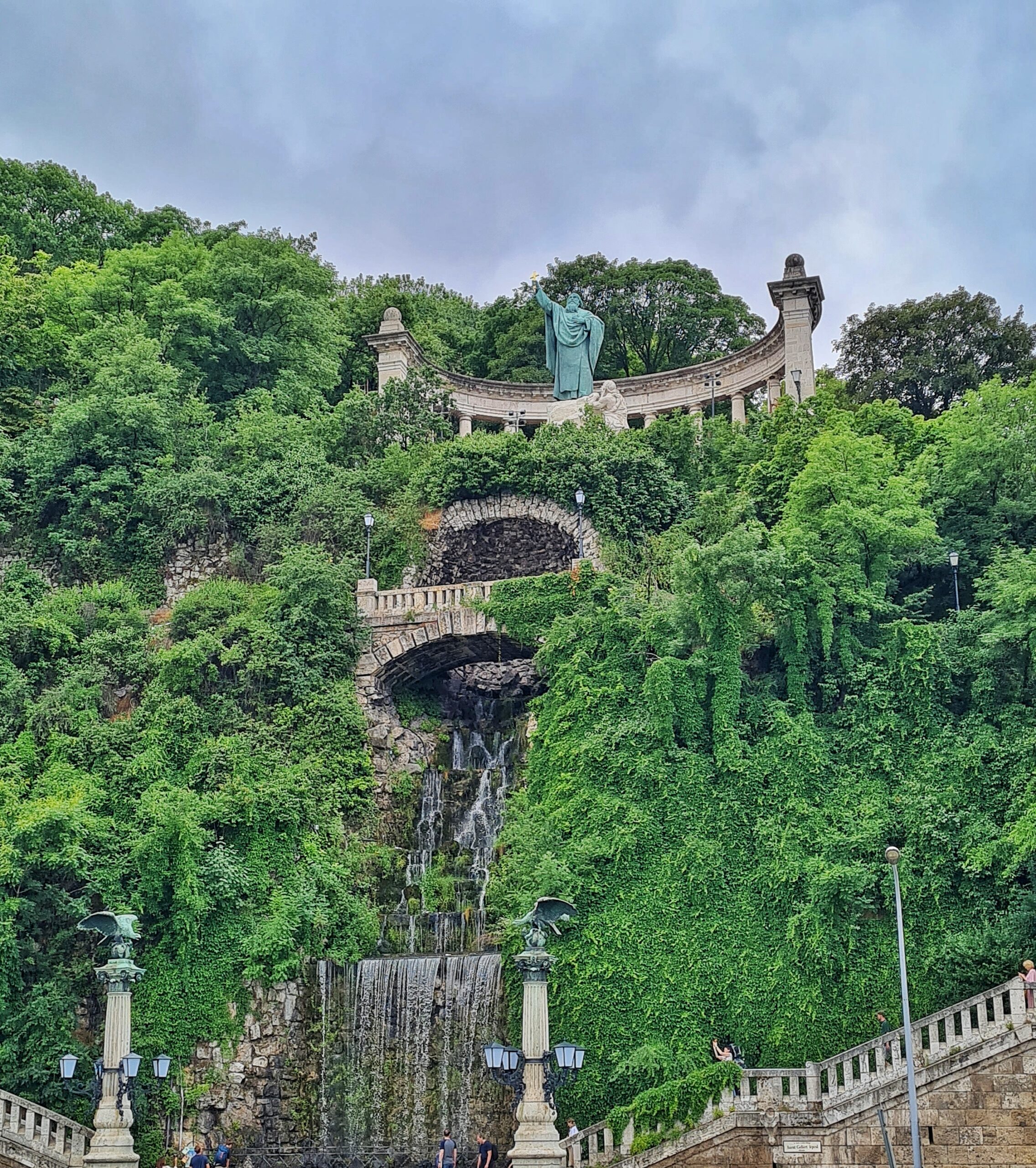 Teil eines Hügels mit einem Wasserfall.´und Bäumen und Büschen. Ganz oben in einer halbkreisförmigen Säulenkolonnade die Statue eines alten Mannes in einem langen Gewand mit Überwurf. Die rechte (aus Sicht der Kamera die linke) Hand hat er erhoben und hält darin ein goldenes Kreuz. Ein Stück darunter eine steinerne Brücke mit Geländer, die über den Wasserfall geht. Und unten zwei steinerne Pfeiler mit Adlern darauf und eine steinerne Terrasse mit Geländer.