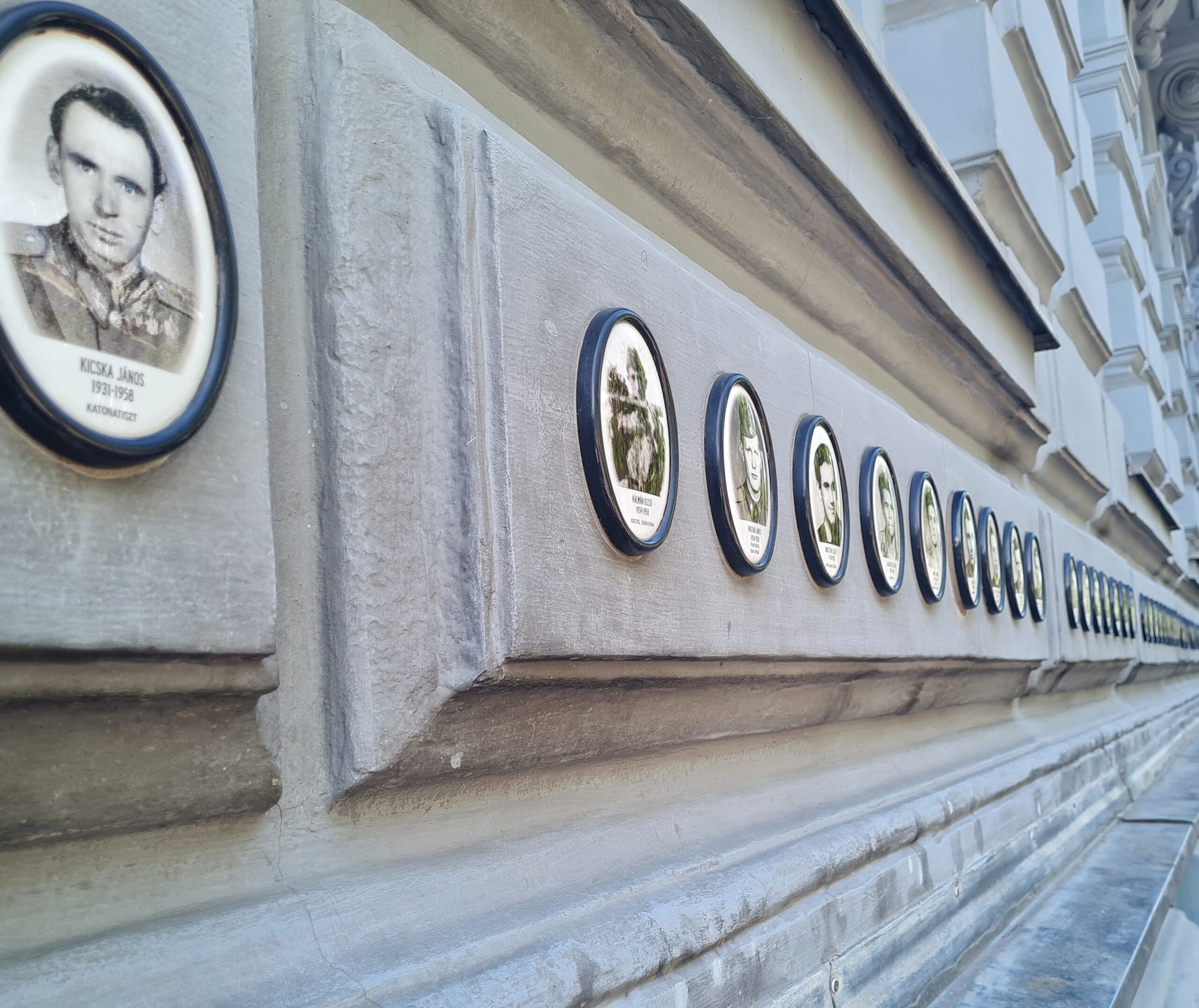 An einer schmuckvollen Hausfassade hängen in runden Rahmen Fotos von einzelnen Menschen mit Text darunter. Das Foto ist seitlich aufgenommen, so dass man die Fotos und den Text nicht genau erkennen kann.