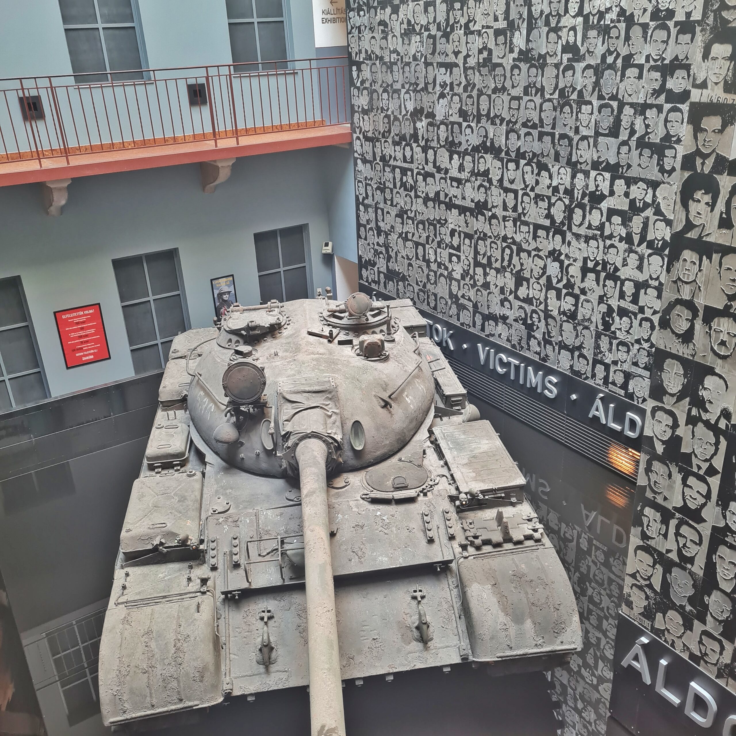 In der Mitte steht ein Panzer. Die hintere Wand ist weiß. mit Fenstern und einem vergitterten Laufgang im ersten Stock. Die rechte Wand ist voller Negativfotos von Menschen. Darunter stehen einige Wörter, von denen man nur das Wirt "Victims" lesen kann.