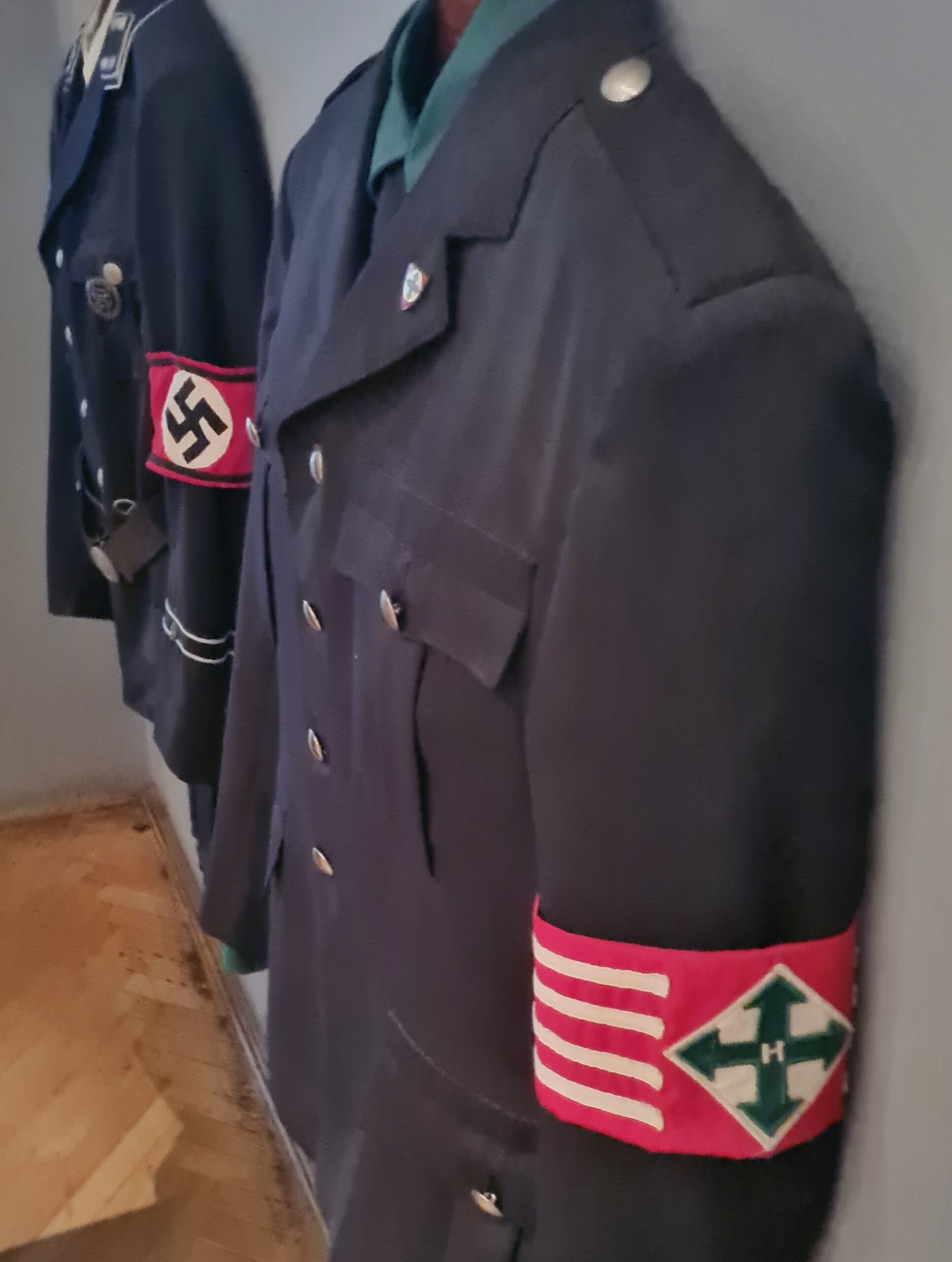 Zwei dunkle Uniformjacken hängen an der Wand. Die hintere hat eine Armbunde mit Hakenkreuz, die vordere eine mit dem Symbol der Pfeilkreuzler (vier Pfeile nach oben, unten, rechts und links; in der Mitte ein H).