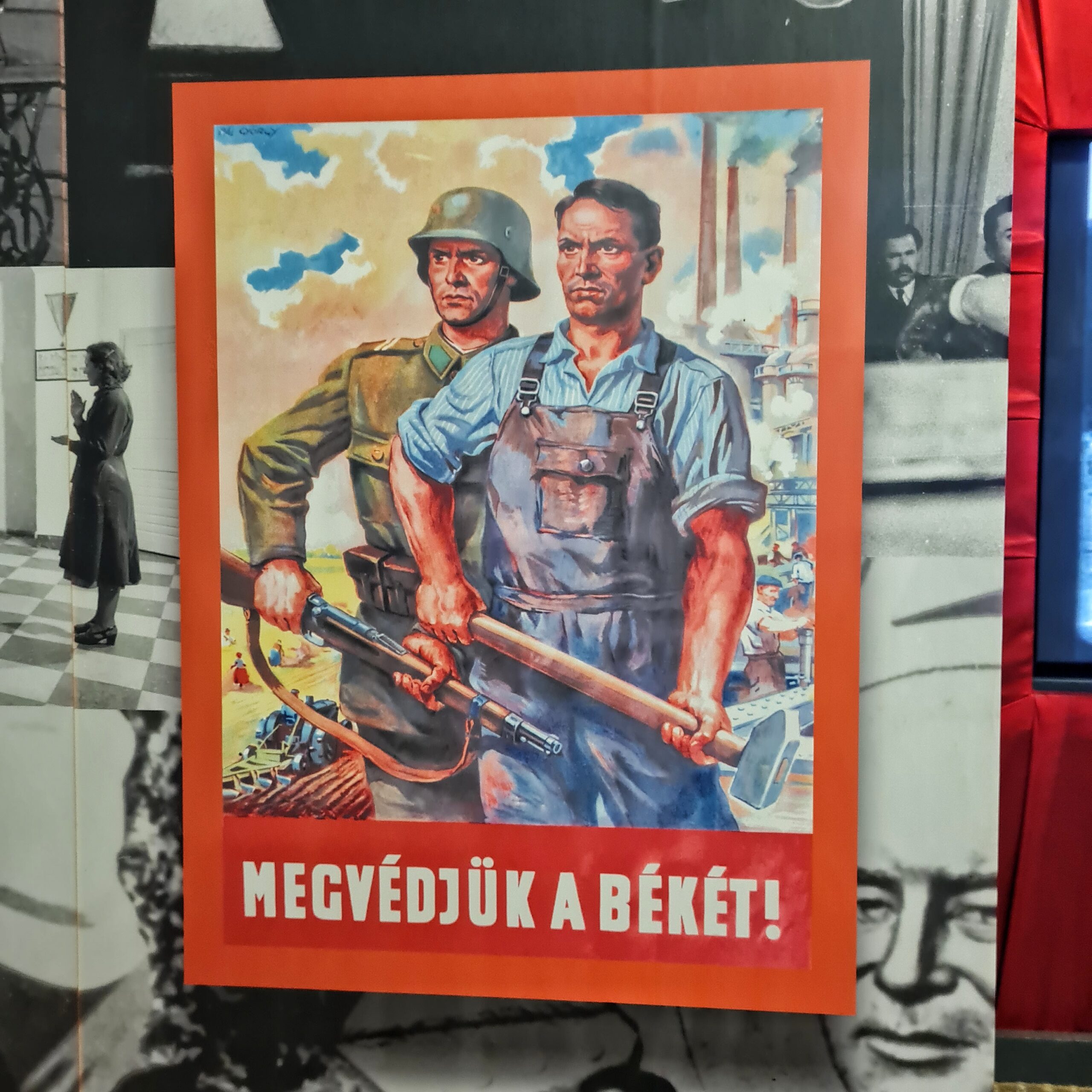 Ein Plakat mit einer Zeichnung, das links einen Soldaten und rechts einen Industriearbeiter zeigt. Der Soldat hält sein Gewehr seitlich, ebenso wie der Arbeiter einen großen Hammer. Im Hintergrund rauchende Fabrikschlote. Darunter eine ungarische Aufschrift.