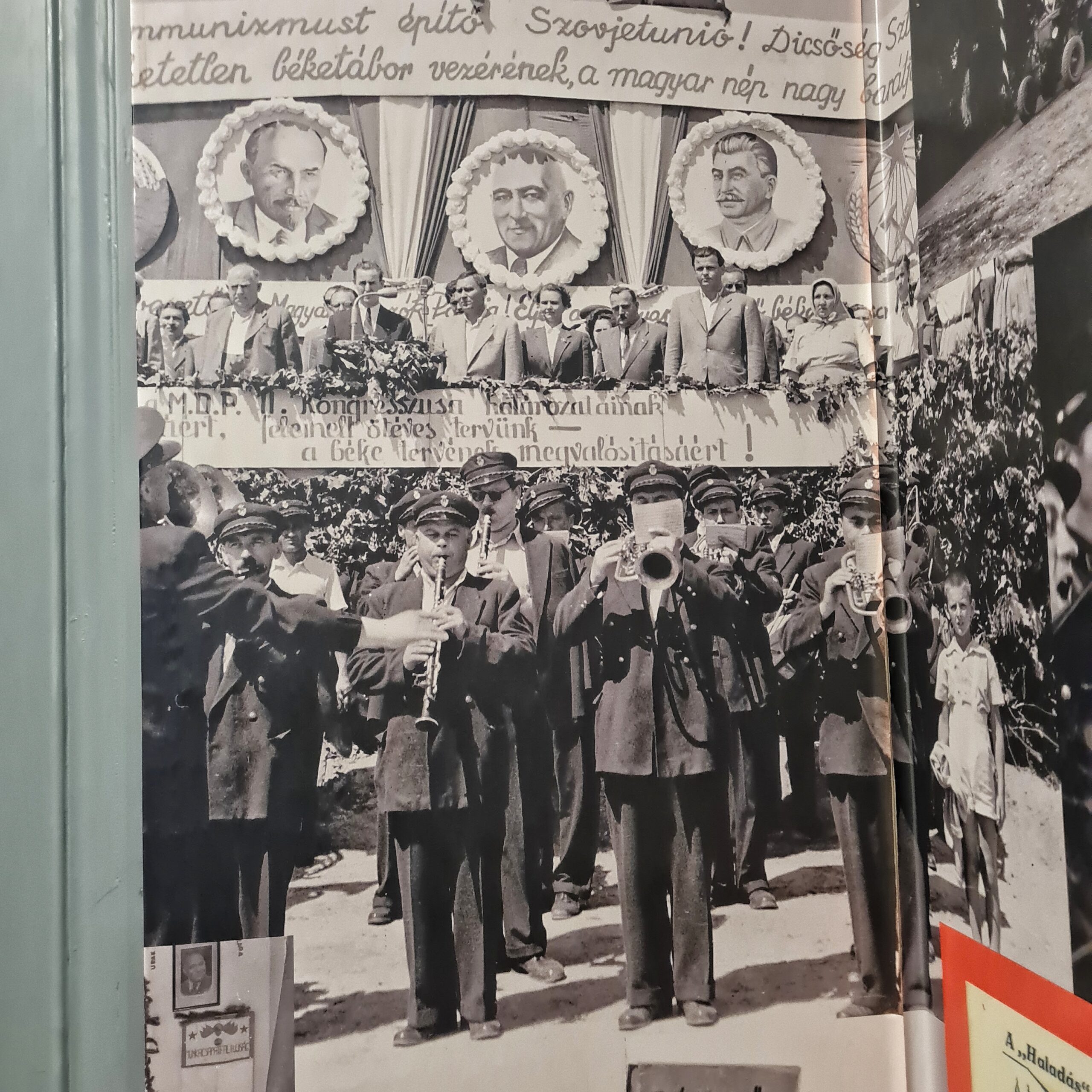 Ein altes Schwarz-Weiß-Foto, das eine Blaskapelle zeigt. Dahinter eine Tribüne, auf der Menschen stehen, und darüber Fotos von Lenin, Stalin und einem weiteren Politiker.