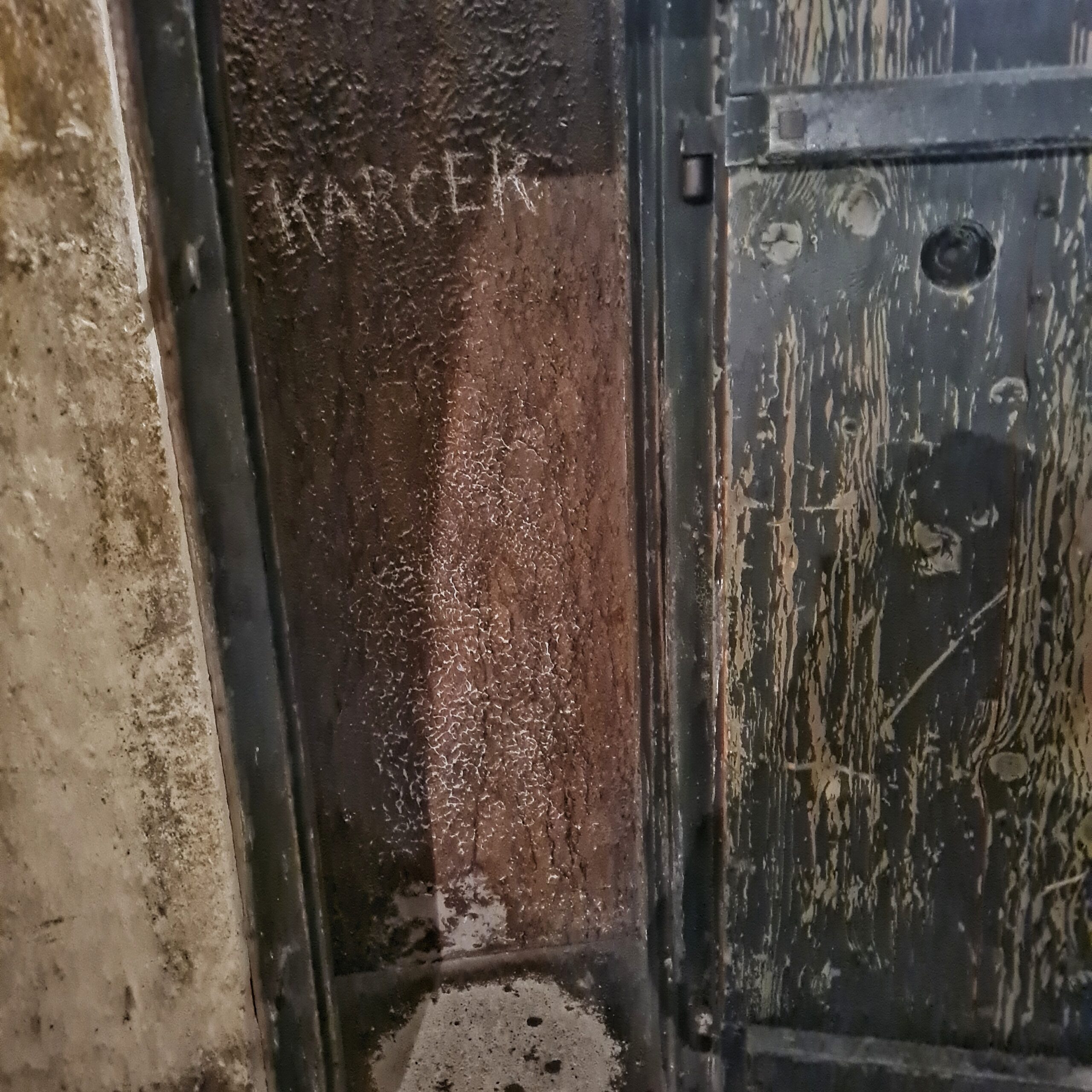 Eine schmale Einbuchtung in einer Metallwand. Darin steht mit Kreide "KARCER". Die große Metalltür davor ist nach außen geöffnet.