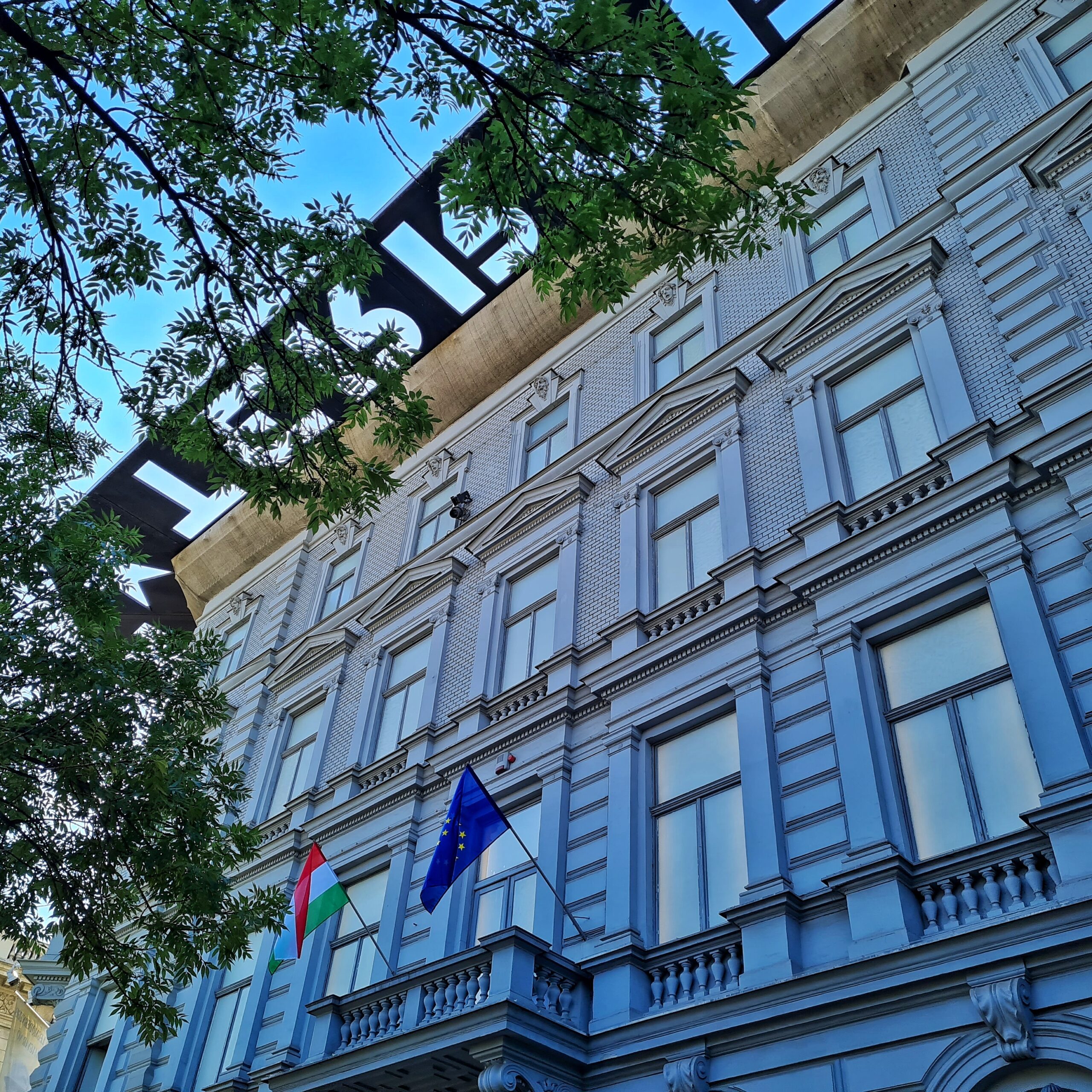 Ein helles, altes Gebäude mit vielen Fenstern, von schräg unten fotografiert. Oben ragt eine Metallplatte nach außen. Ihre Aussparungen bilden Buchstaben. Unten hängen über einem Balkon die ungarische und die europäische Flagge.