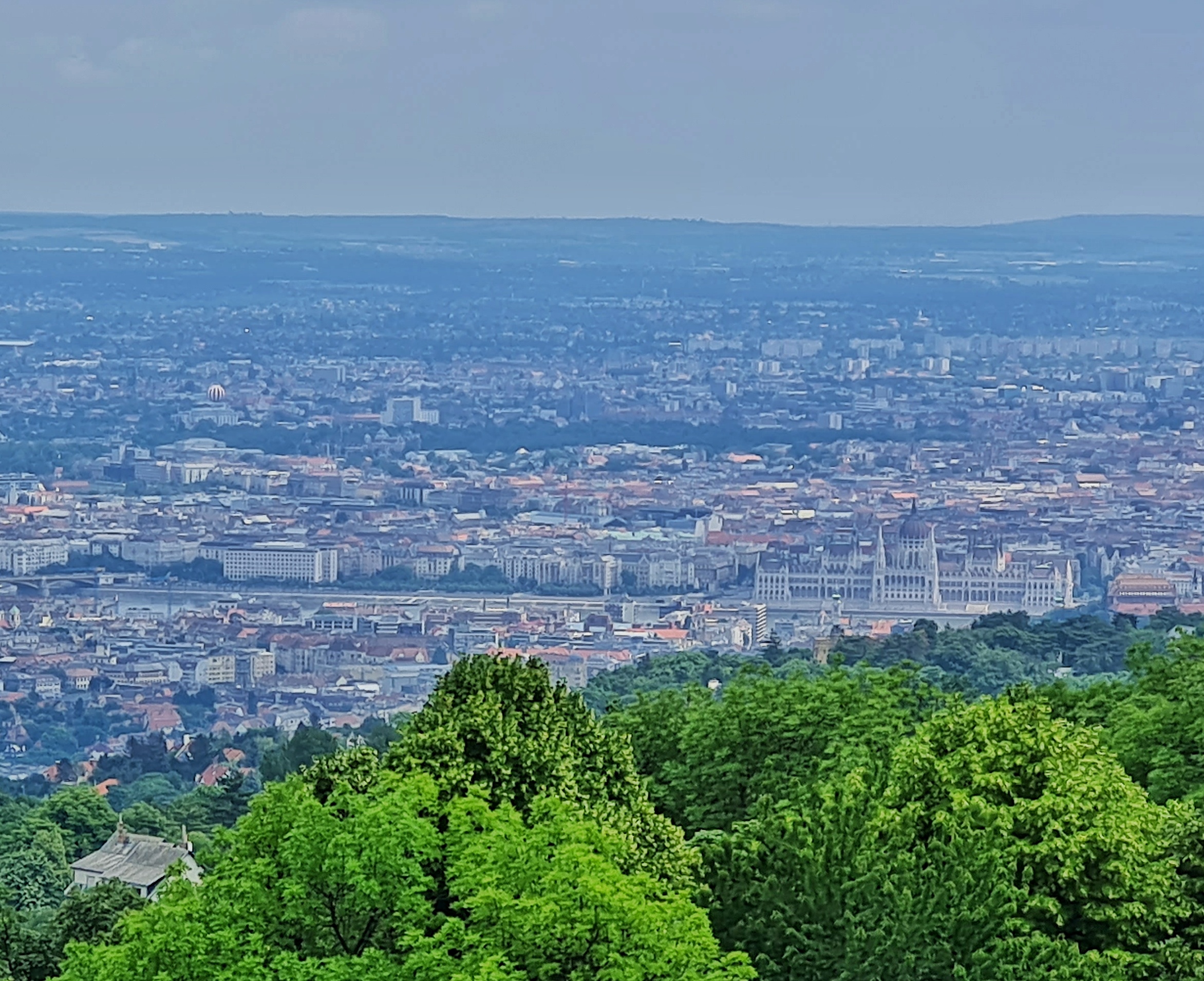 Blick auf eine weite Ebene mit Bebauung. Man erkennt undeutlich das Parlametnsgebäude. Im Vordergrund einige Bäume.