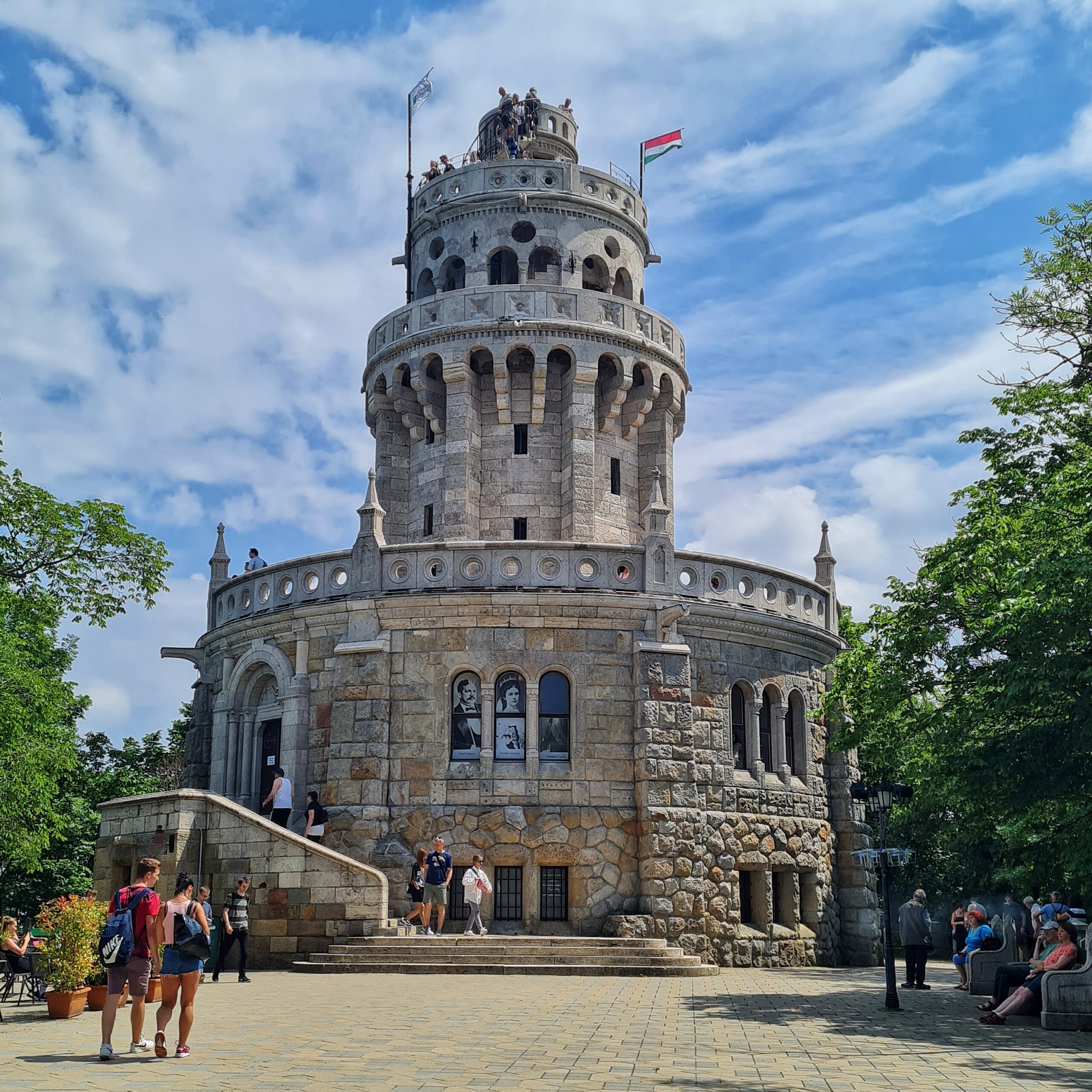 Ein Turm aus hellen Steinen. Unten ist er sehr breit. Die Ebene darunter ist deutlich schmaler. Darüber ist eine etwas schmalere Ebene; am Rand weht die ungarische Flagge. Ganz oben ist ein noch schmaleres Türmchen. Vor und auf dem Turm sind einige Menschen.
