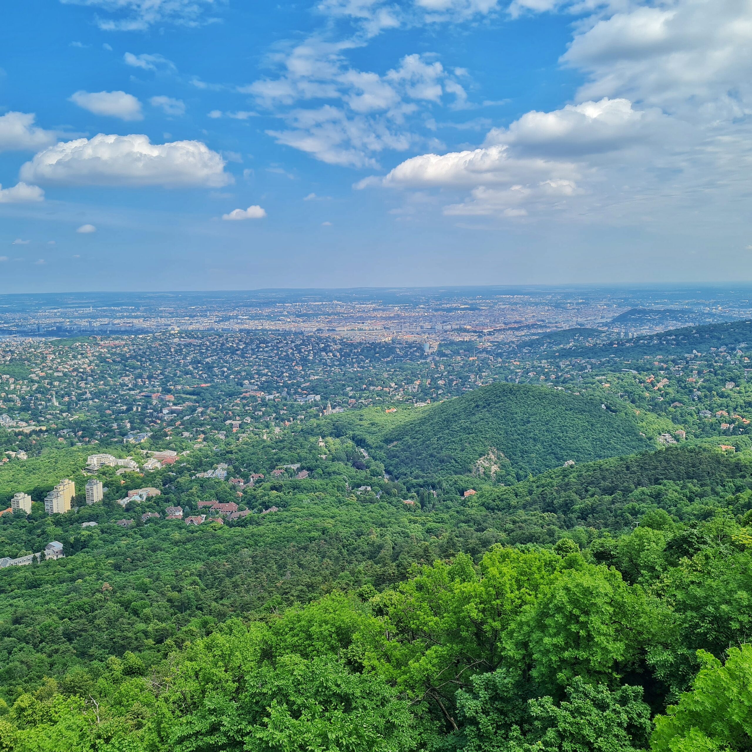 Blick auf grüne Hügel (teilweise mit Häusern) und im Hintergrund eine weite Ebene. Der Himmel darüber ist hellblau mit einigen Wolken.