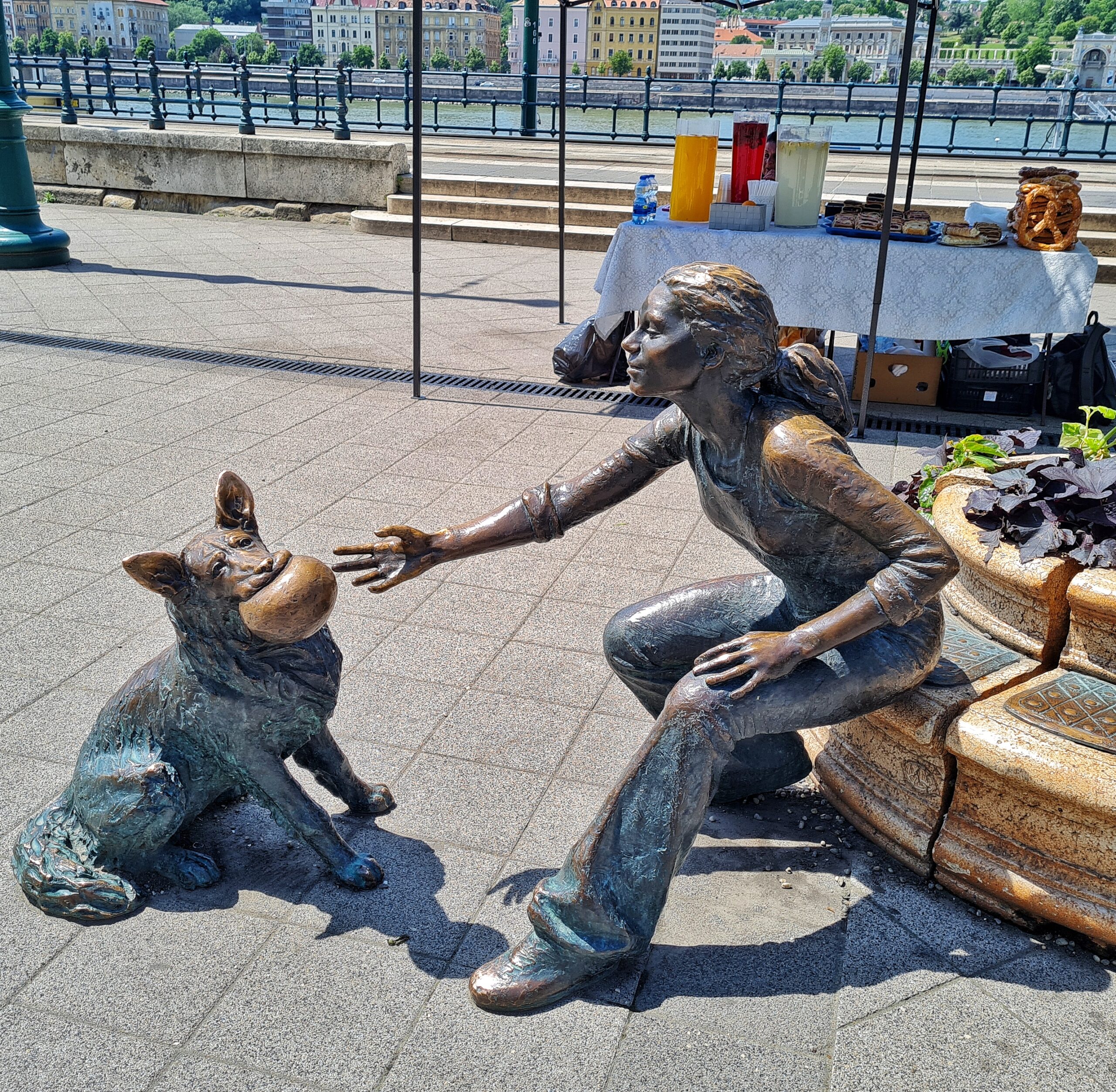 Auf einer gepflasterten Fläche die Statuen eines Mädchens und eines Hundes. Der Hund hat einen großen Ball im Maul. Dass Mädchen mit Pferdeschwanz und langen Hosen sitzt rechts von ihm und greift mit einer Hand nach dem Ball. Dahinter ein Stand mit Getränken und Kuchen. Dahinter der Fluß und die Gebäude auf der anderen Uferseite.