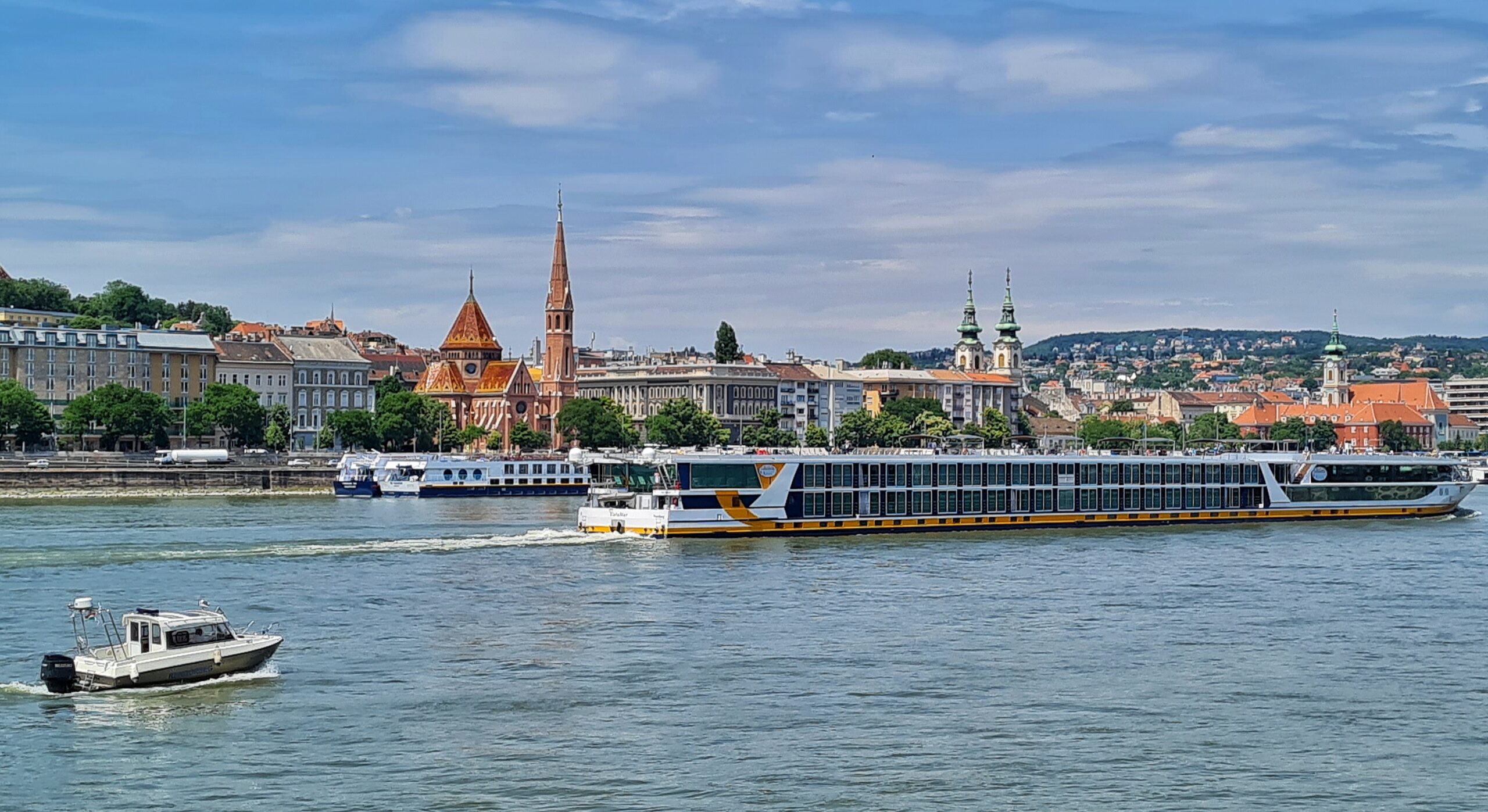 Blick über den Fluß auf eine Kirche mit spitzen Turm und hohe Gebäude, rechts mit zwei Türmen. Auf dem Fluß fährt eine flaches zweistöckiges Schiff mit dicht an dicht liegenden hohen Fenstern. Am anderen ufer leigen weitere Schiffe.