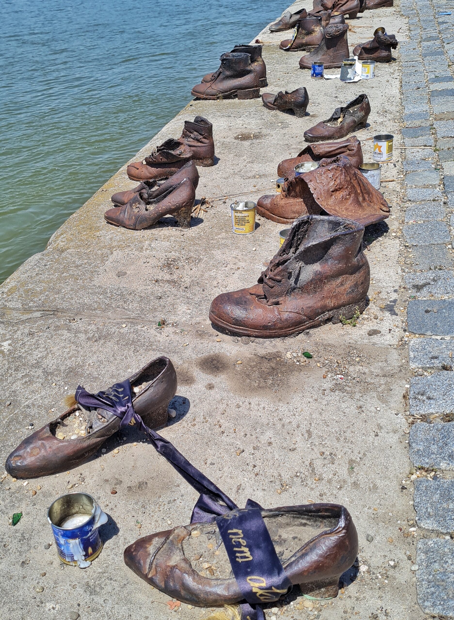 Mehrere Metallschuhe, die am Rand des Flußes stehen. Sie sind völlig unterschiedlich: Damen schuhe mit Absatz, ausgelatschte Stiefel, Halbschuhe, alles in unterschiedlichen Größen. Neben einige Schuhen stehen Metalldosen mit Kerzenwachs darin. Die beiden vordersten Schuhe sind mit einem breiten blauen Band mit einer nicht erkennbaren Inschrift verbunden.