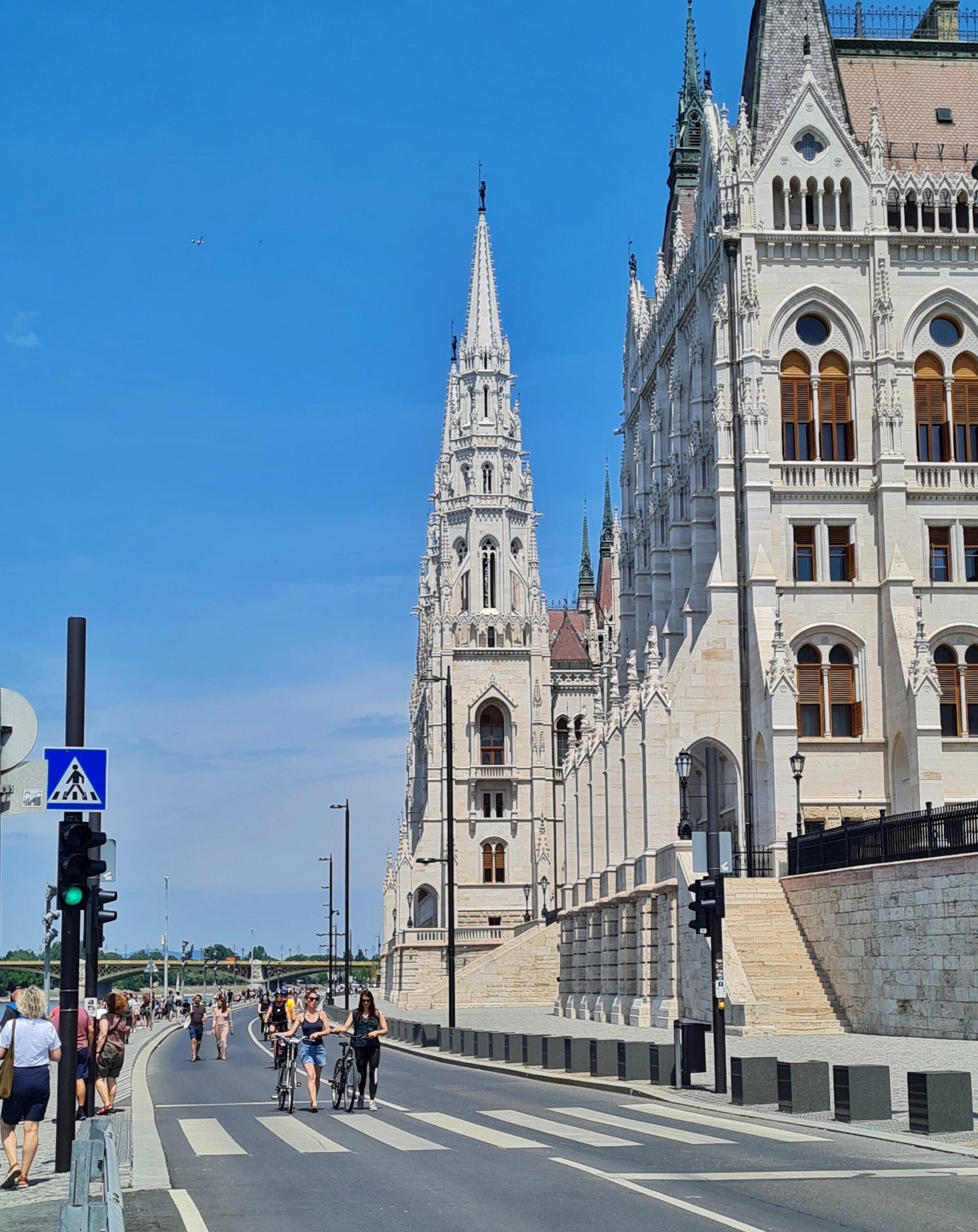 Das gleiche Gebäude seitlivh aus einiger Entfernung fotografiert. Man sieht links davon die Straße mit einer Ampel und einem Zebrastreifen, auf der Menschen zu sehen sind, die fahrradfahren oder ihr Fahrrad schieben. Außerdem gehen dort auch Menschen, ebenso auf dem Gehweg ganz links im Bild.
