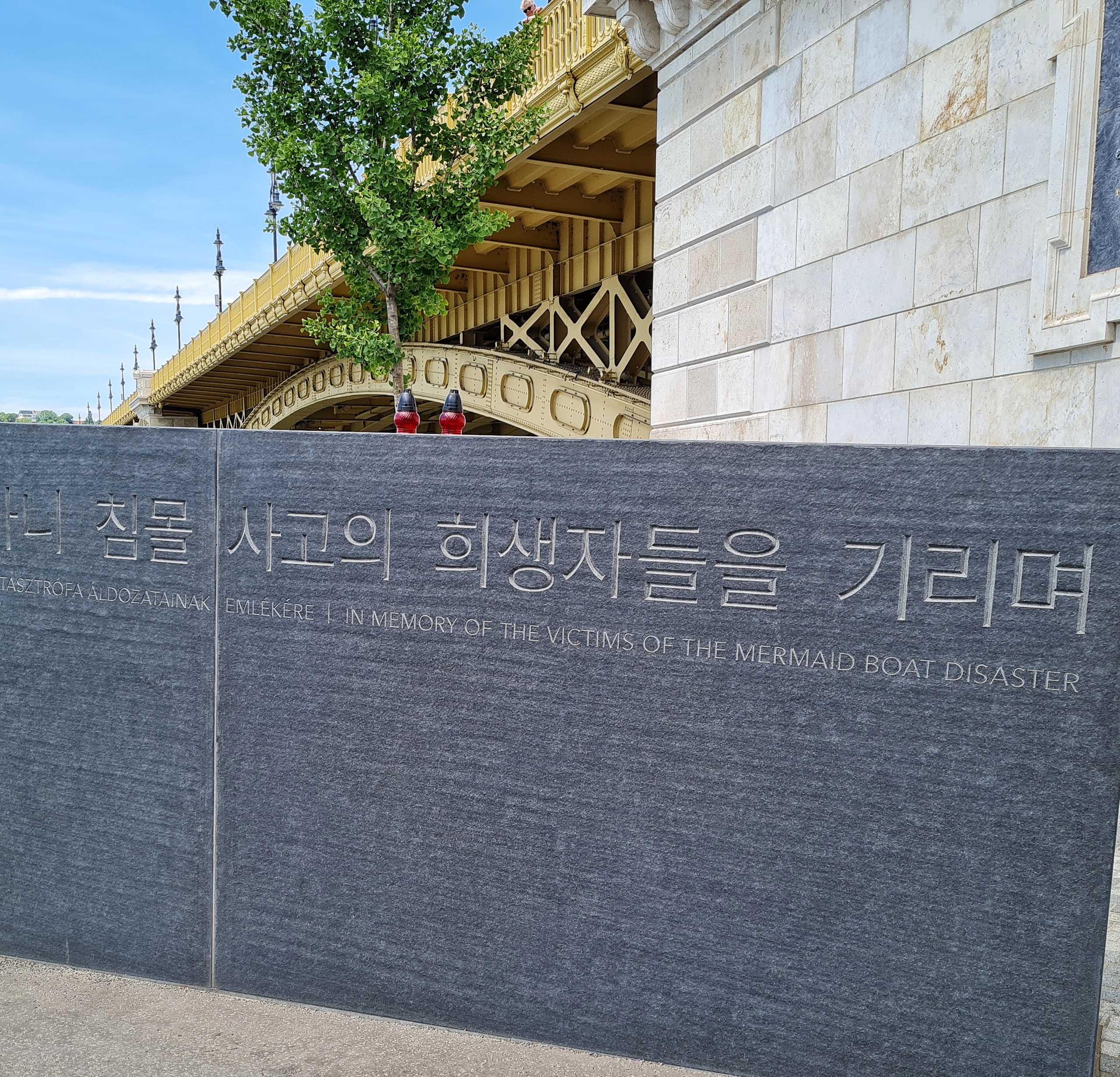 Eine große dunkelgraue Steinplatte, die aufrecht vor einem Brückenpfeiler steht., Darauf Schriftzeichen und die englische Aufschrift: "In Memory of the Victims of the Mermaid Boat Disaster".