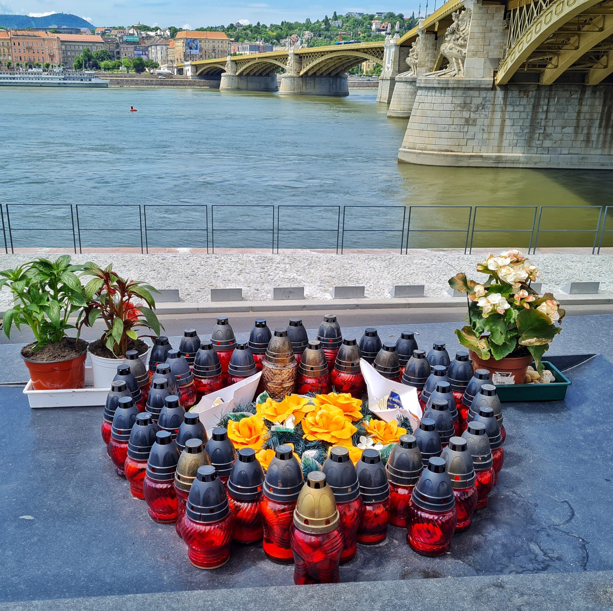 Blick auf den Fluß und die Brücke am rechten Rand des Fotos. Im Vordergrund viele rote Grabkerzen, die gelbe Rosen einrahmen. Links und rechts stehen kleine Blumentöpfe mit blühenden Pflanzen.