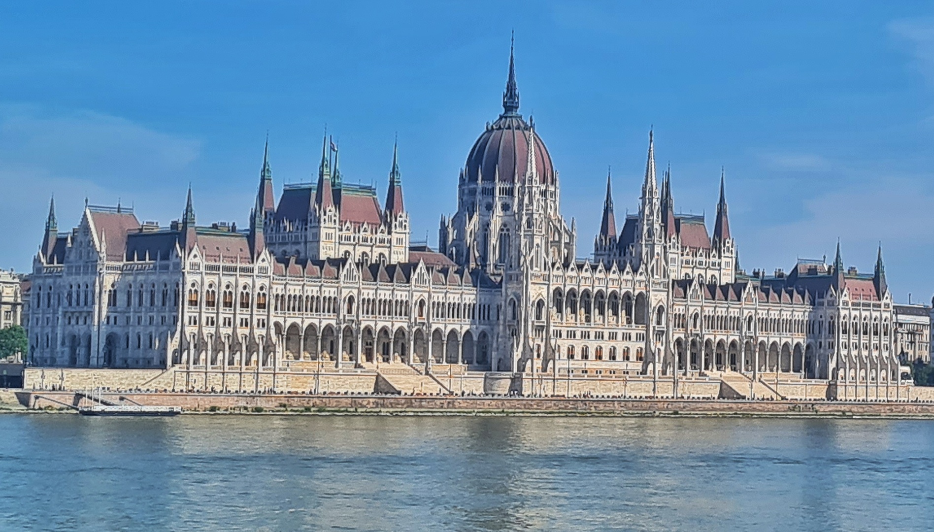 Ein großes, helles Gebäude mit vielen spitzen Türmchen. In der Mitte ein runder Turm mit einer roten Kuppel und einem spitzen Turm darauf. Links ein viereckiges rotes Dach. An den Ecken spitze Türmchen mit roten Dächern. Rechts ist ein Teil eines gleichen Daches zu sehen, Unten links gehen einige Stufen zu einem Säulengang empor. Davor ein Fluß.