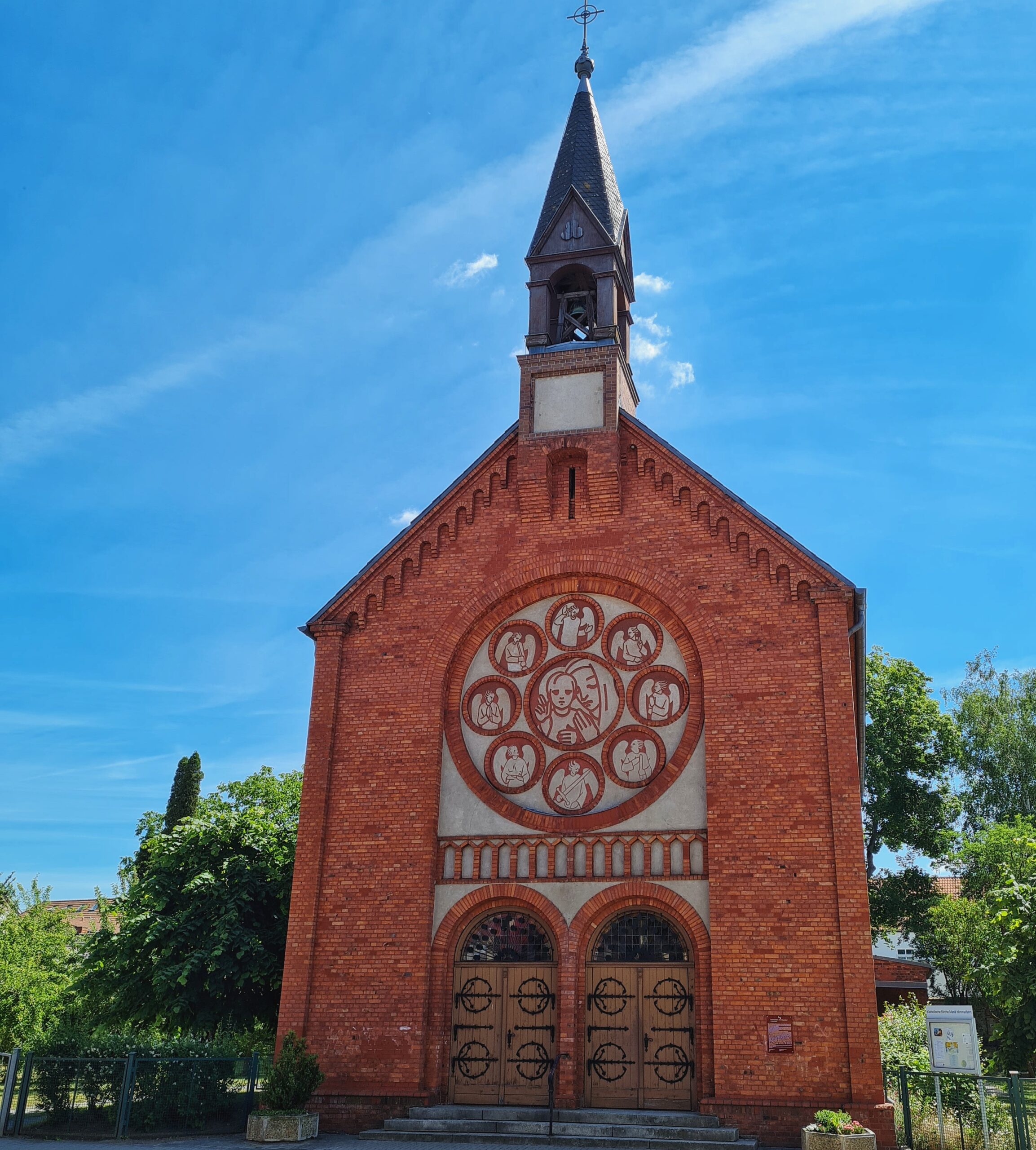 Eine kleine Kirche aus rötlichem Backstein von vorne fotografiert. Die beiden großen Eingangstüren aus Holz mit schwarzen Verzierungen und halbrundem Glas darüber sind geschlossen. Darüber ist ein großes rundes Ornament. Dieses zeigt in der Mitte in einem Kreis eine Frau mit einem Kopftuch, die schützend ihre Hände über ein Kind hält. Außen sind acht kleinere Kreise, die verschiedene Engelsdarstellungen zeigen. Ganz oben ist ein kleiner spitzer Kirchturm.