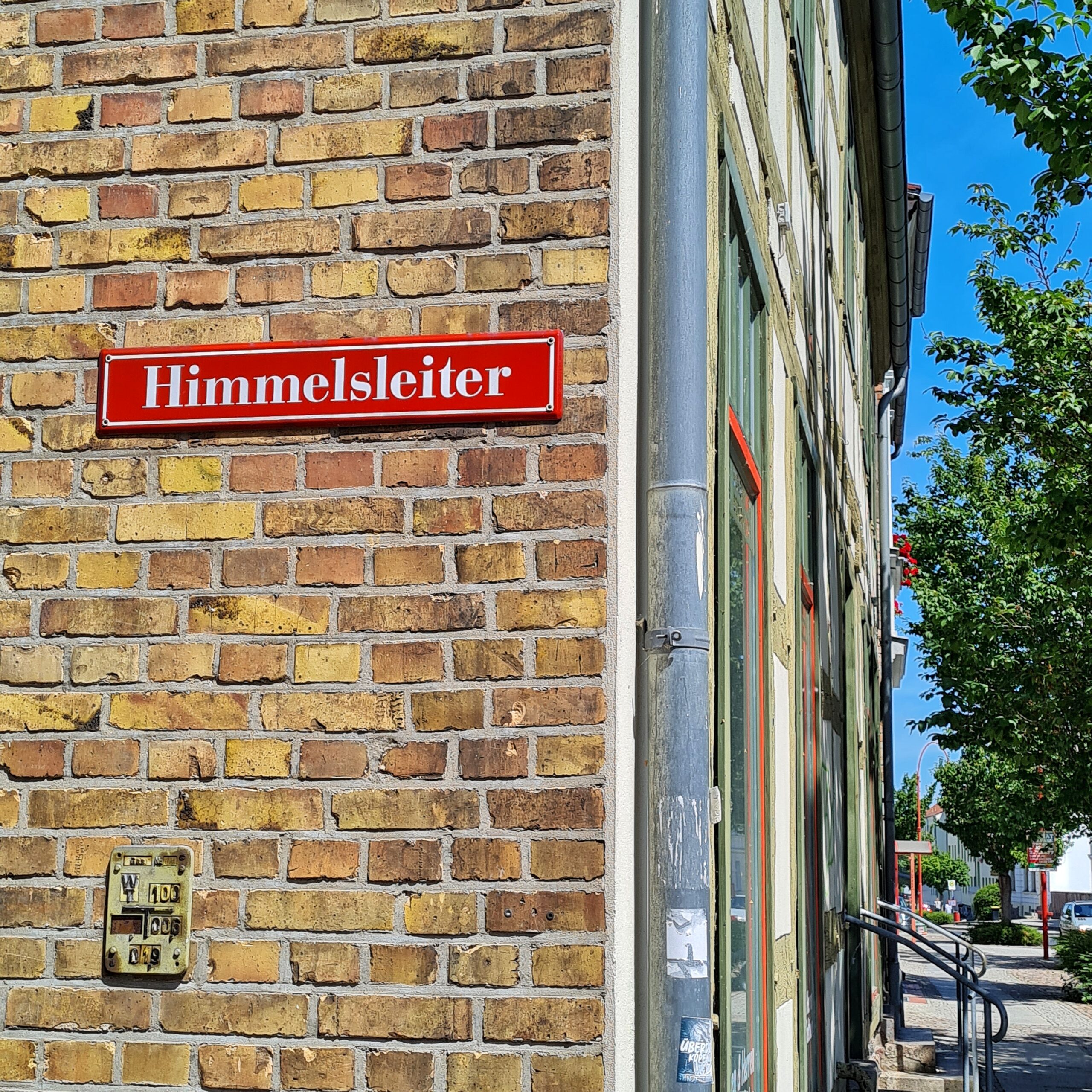 Eine Hausecke. Rechts ist die Front eines kleiinen Ladens und ein paar Stufen mit Geländer zu erkennen,maußerdem ein Gehweg und Bäume. Das linke Drittel des Fotos zeigt eine gelbe Backsteinmauer, an der ein rotes Straßenschuld angebracht ist. In weißer Schrift steht darauf: Himmelsleiter.
