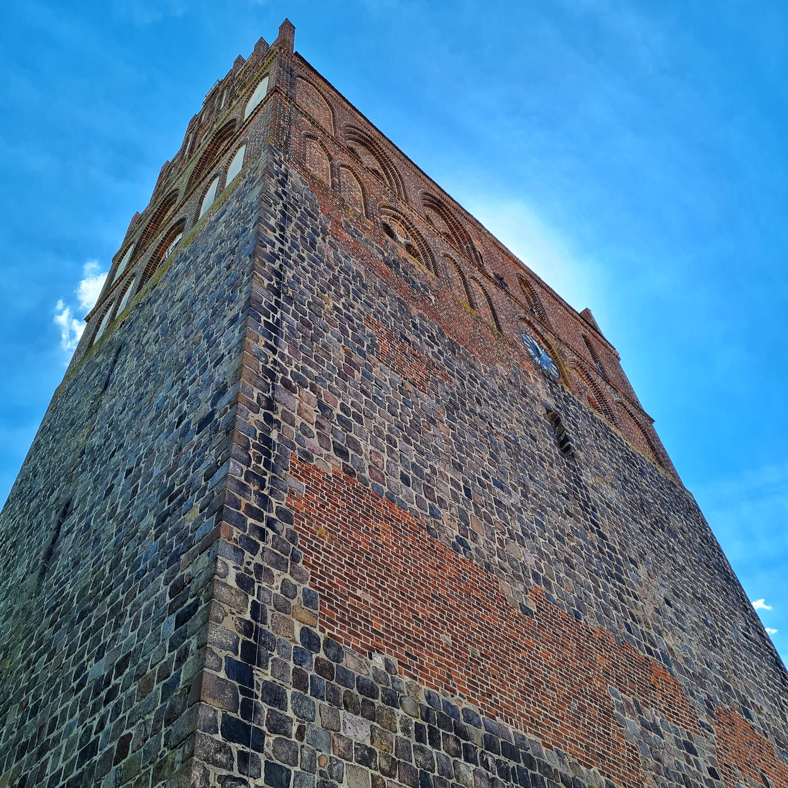 Der Kirchurm aus rotem Backstein von unten nach oben fotografiert. Er ist hoch und rechteckig mit einer sehr breiten Seite und wirkt daher massiv und wuchtig.