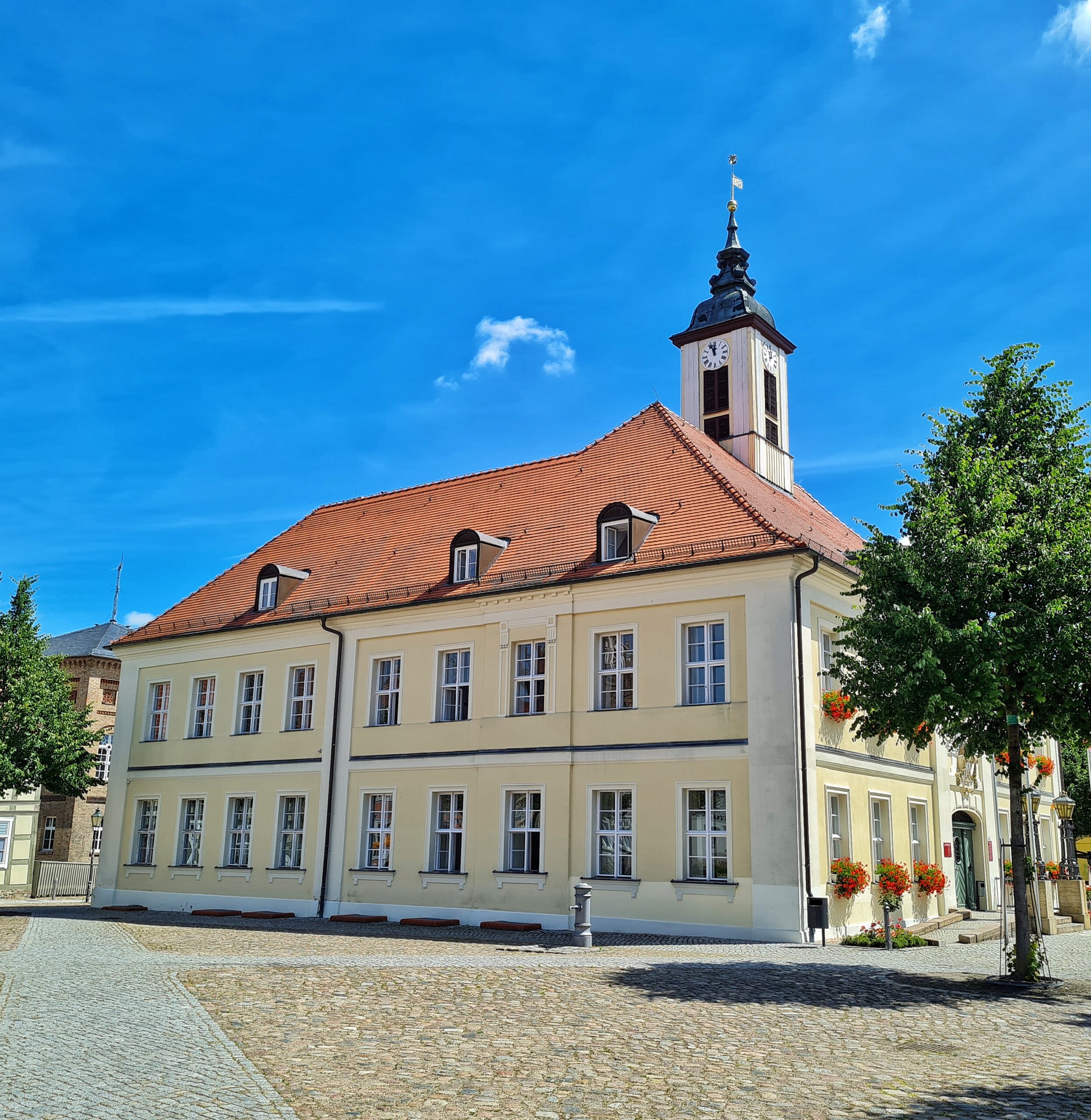 Ein viereckiges Gebäude auf einem gepflasterten Platz. Es ist in einem hellen gelblichen Ton gestrichen, wobei die Ränder weiß sind. Das Foto wurde schräg-seitlich aufgenommen. Dadurch sieht man eine Seiten des Gebäudes vollständig. Diese besteht aus 18 weißen Sprossenfenstern, neun im Erdgeschoß und neun im Obergeschoß. Im roten Dach sind drei Gauben. Rechts sieht man den Eingangsbereich mit einer geschlossenen Tür und weiteren Sprossenfenstern, vor denen Blumenkästen mit leuchtend-roten Blumen hängen. Oben ist ein kleiner weißer Turm. Links und rechts vom Gebäude stehen mittelgroße Bäume.
