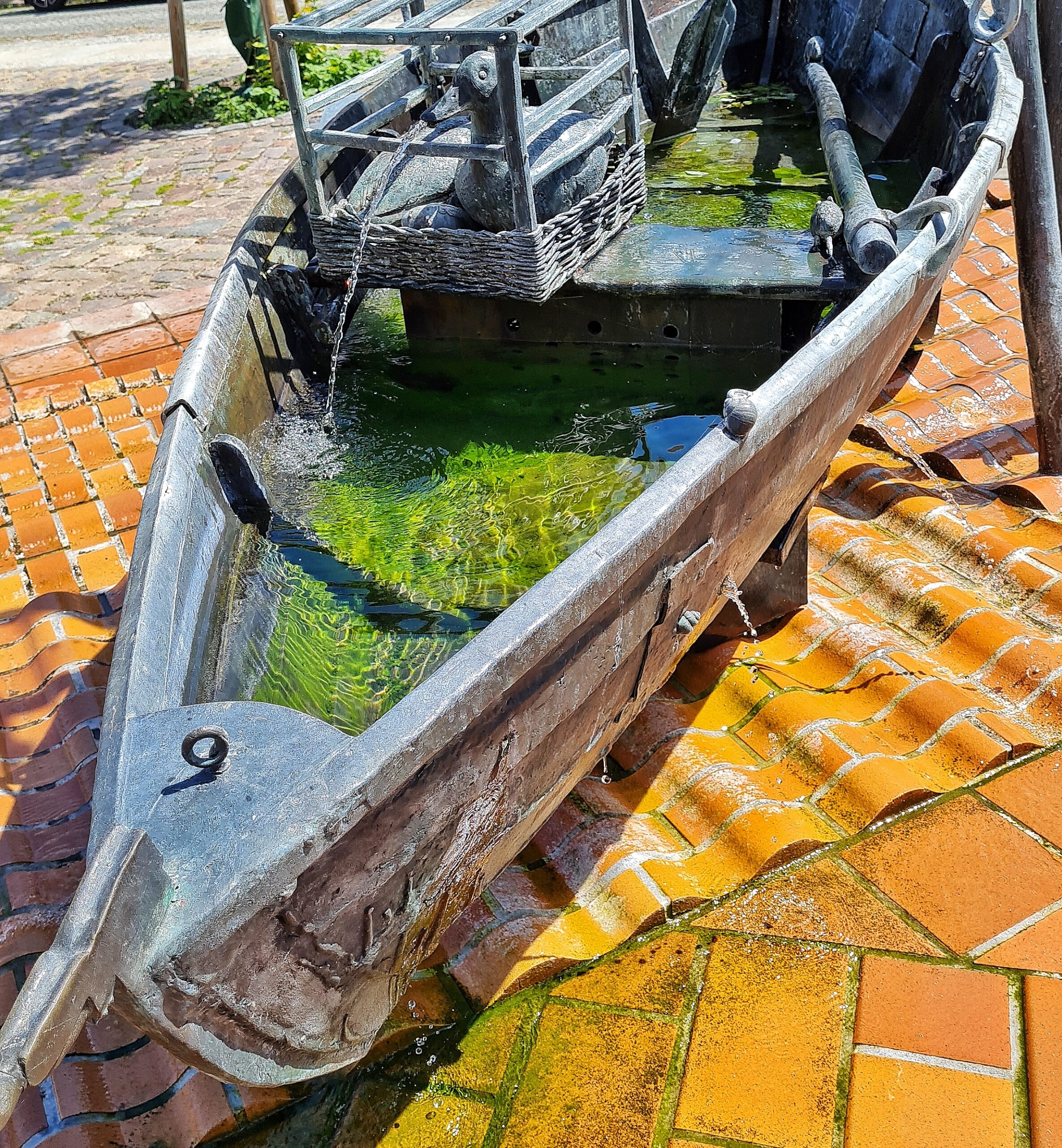Ein großes Ruderboot aus Metall. Im Boot ist Wasser. Auf der Bank in der Mitte des Bootes steht ein viereckiger Metallkorb mit zwei Metallenten darin. Die Ente, die in Richtung Kamera schaut, speit Wasser aus ihrem Schnabel.