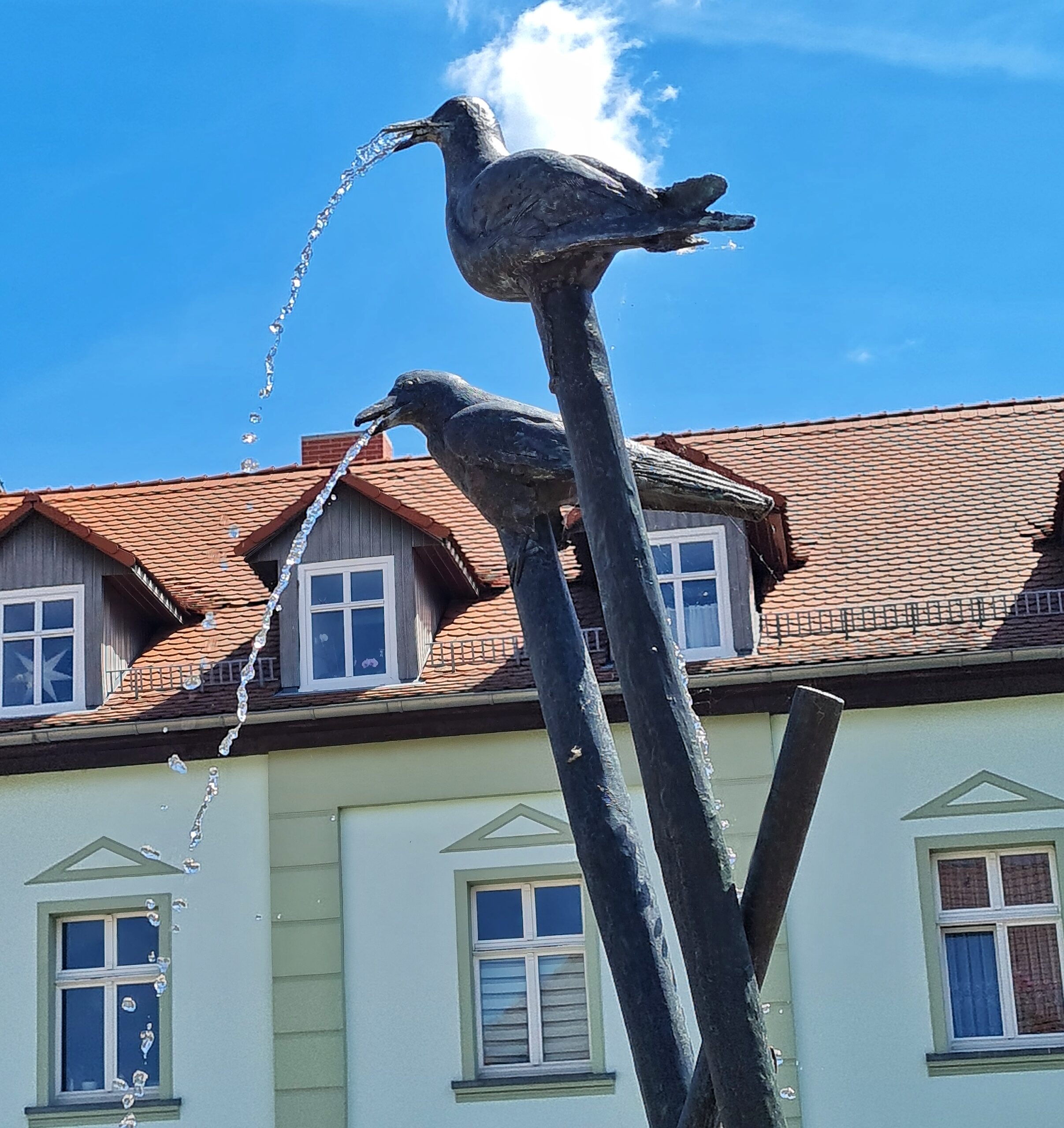 Im Hintergrund der obere Teil eines hellen Hauses mit Fenstern und mit Dachgauben im roten Dach. Im Vordergrund zwei Metallpfosten, auf denen Metallvögel hocken. Aus ihren Schnäbeln spritzt Wasser.