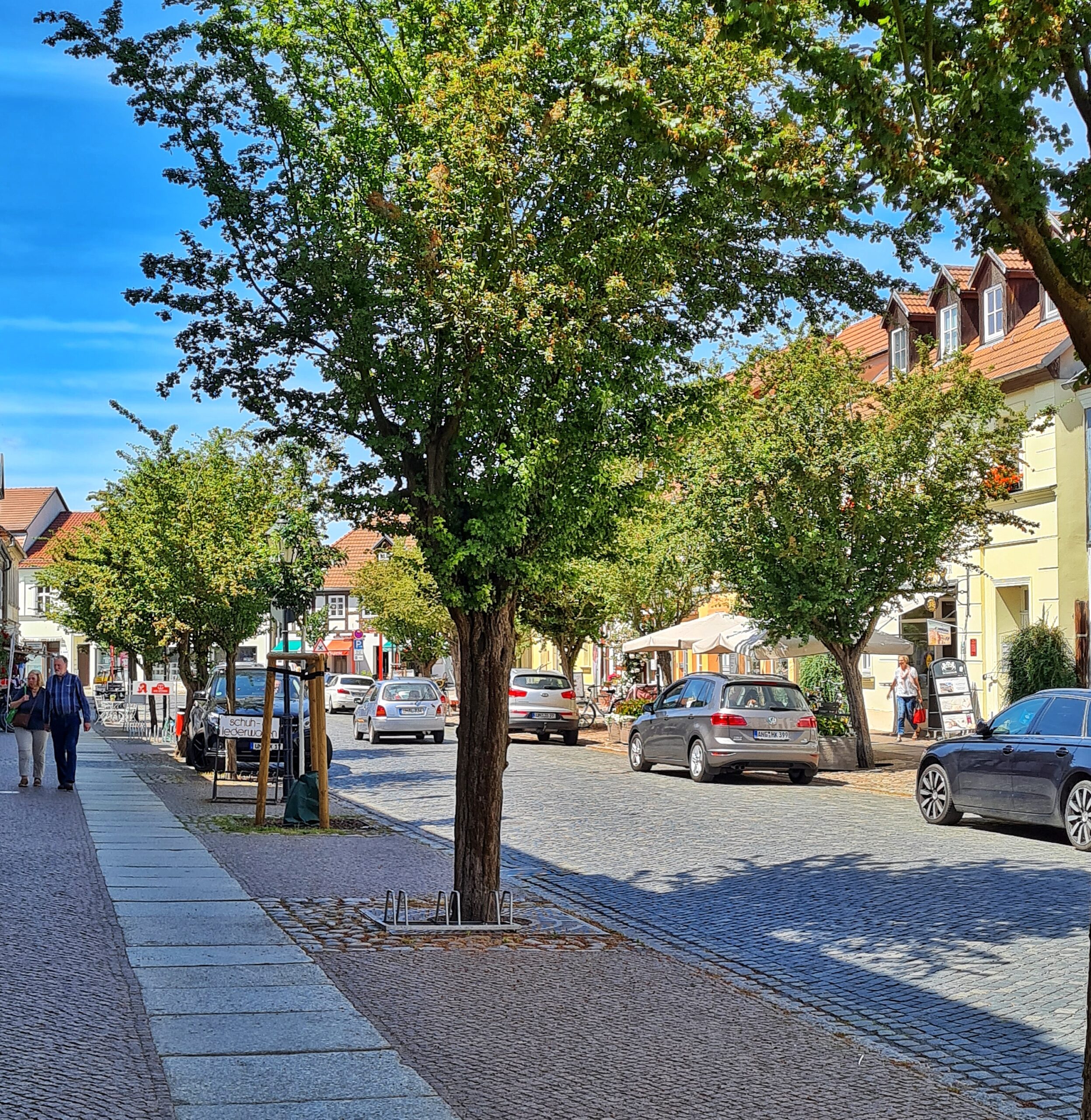 Eien gepflasterte Straße mit breiten Gehwegen. Mittelgroße belaubte Bäume am Straßenrand werfen Schatten. Auf der Straße fahren und parken einige Pkw. Auf den Gehwegen vereinzelt Fußgänger. Es sind Schilder von Geschäften zu sehen und auf der rechten Straßenseite ein großer viereckiger Sonnenschirm.