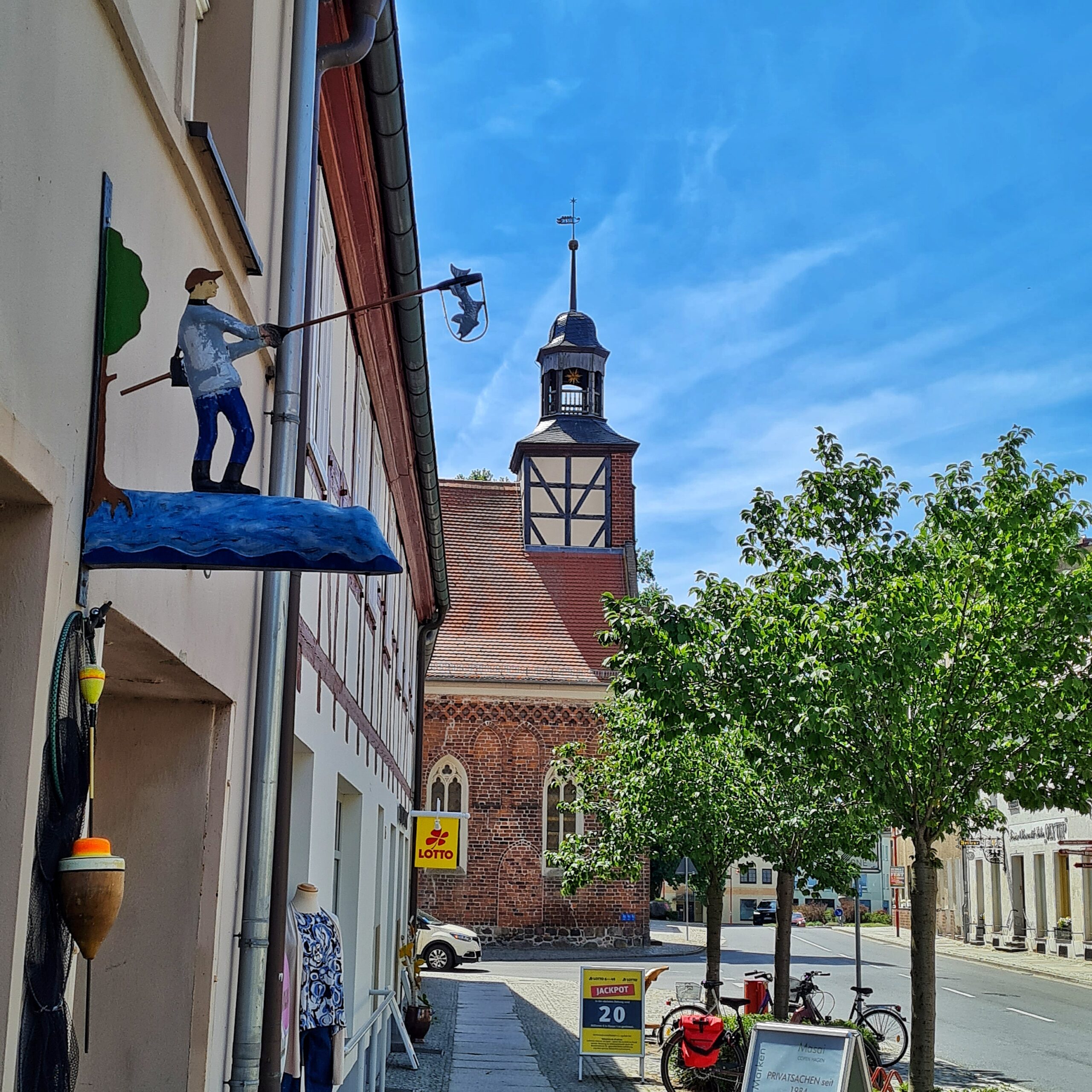 Blick entlang einer Häuserfront auf eine kleine Krche, von der man nur einen Teil mit einem kleinen Turm sieht. Rechts eine Straße, ein paar kleine Bäum,enund ein Gehweg, an dessen Rand Fahrräder und Reklameaufsteller stehen. Die Häuserfront besteht aus zwei Gebäuden. Das hintere mit Fachwerk. Am vorderen ragt etwas in die Luft: ein blauer Untergrund, der aussieht wie Wasser. Zum Haus hin ein halber Laubbbaum. Auf dem Blauen steht eine männliche Figur, die eine Angel mit einem Fisch daran in die Luft hält.