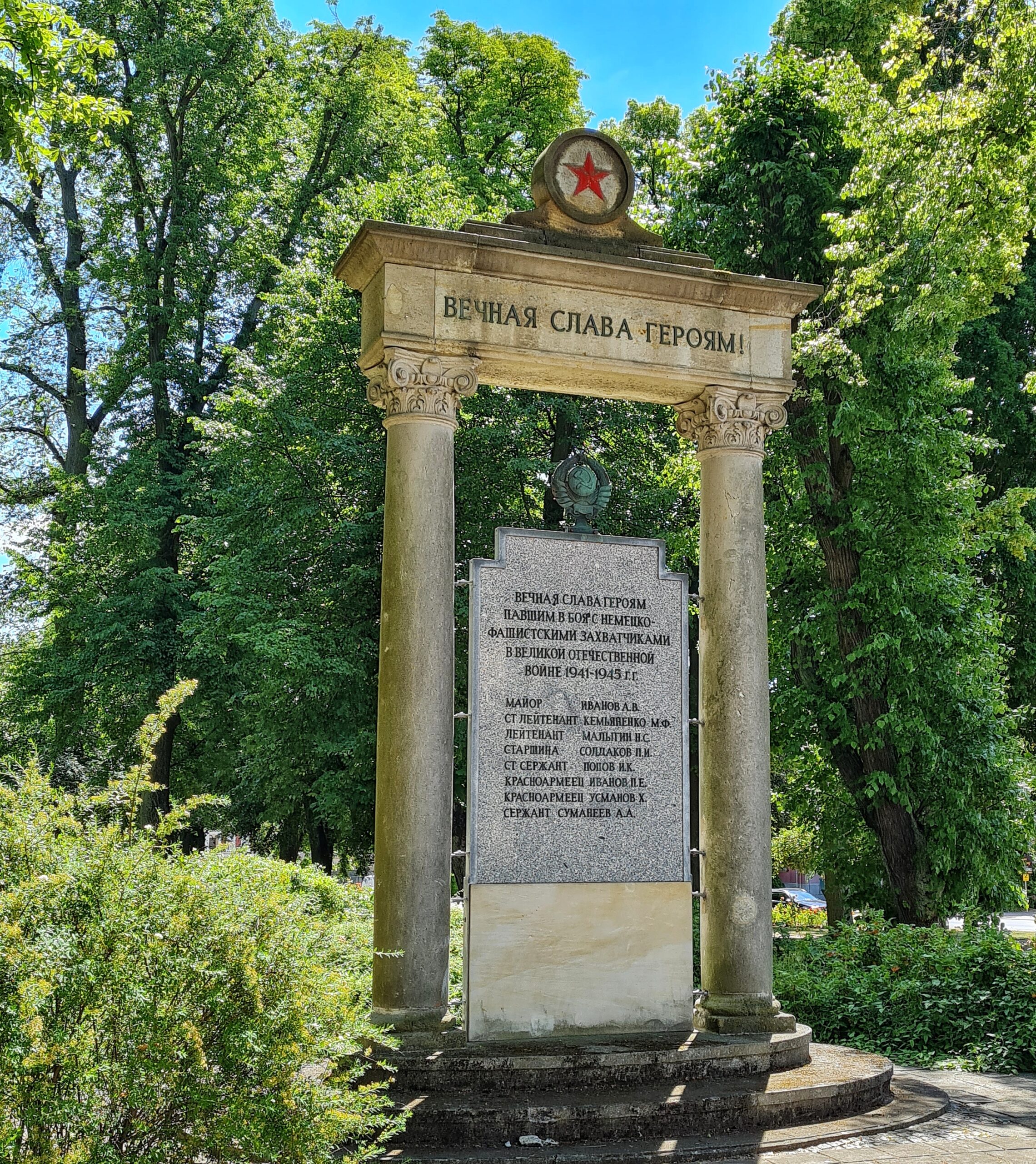 Ein kleiner Park mit Bäumen. Darin ein Steinportal, oben mit einer russischen Inschrift. Darüber eine kreisförmige Spitze mit einem roten Stern darin. Im Portal steht eien grabsteinähnliche Platte mit Text darauf.