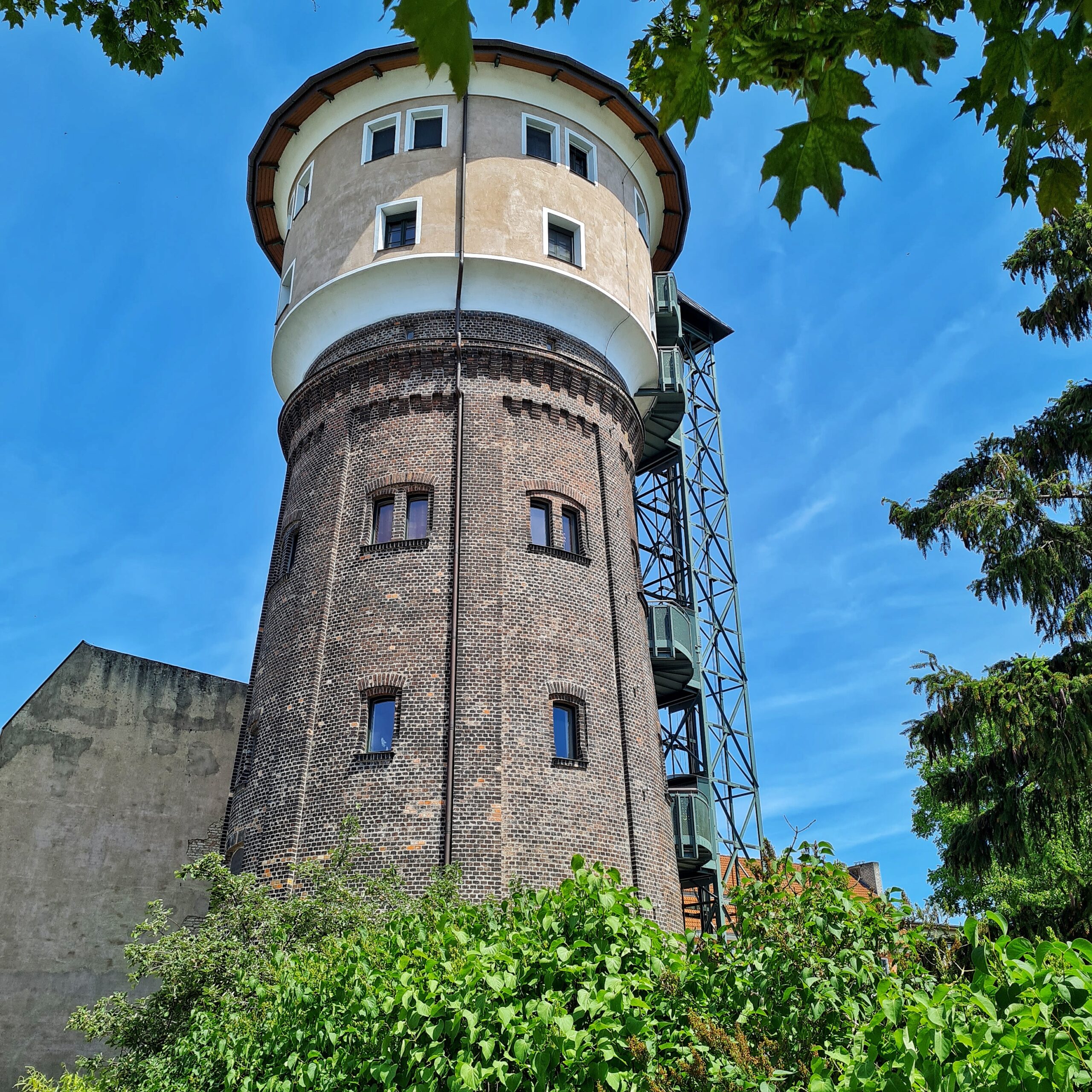 Ein Wasserturm mit mehreren kleinen Fenstern. Der untere Teil ist dunkelgrau, der obere hell. An der rechten Seite ist außen ein Metallgestell. Unten sind einige Büsche, und oben und rechts ragen Blätter ins Foto.