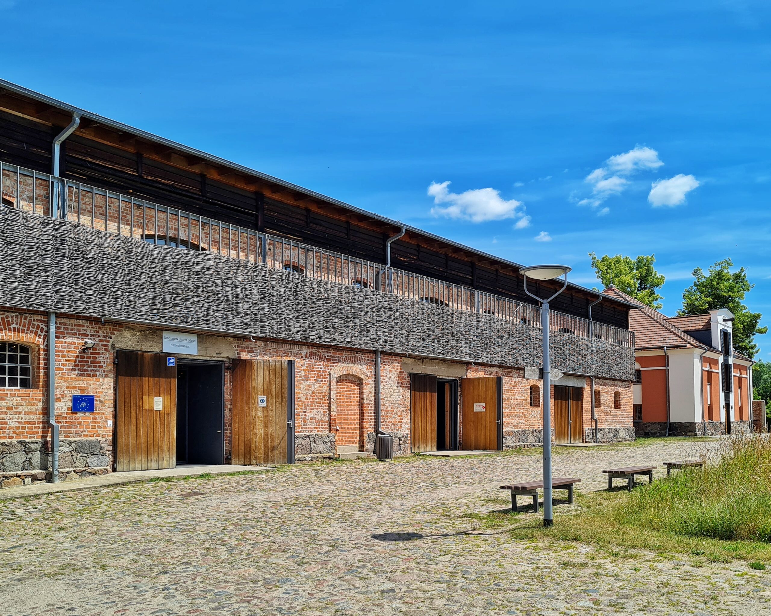 Ein niedriges langes Gebäude. Oben sieht man die oberen Kanten von abgerundeten Fenstern. Die Fenster selbst sind verdeckt durch eine Art Spalier mit Stroh. Im Erdgeschoß drei breite Doppeltüren aus braunem Holz, von denen jeweils ein Flügel nach außen geöffnet ist.