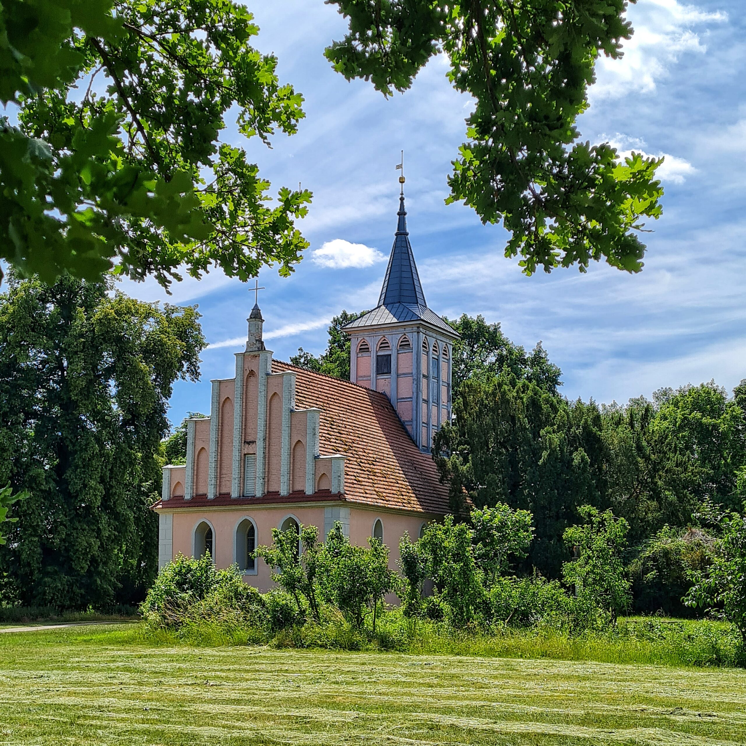 Hiner einer gepflegten Rasenfläche, umstanden von hohen Bäumen eine Kirche mit einem Fachwerkturm und einem hellrosa Kirchenbau.