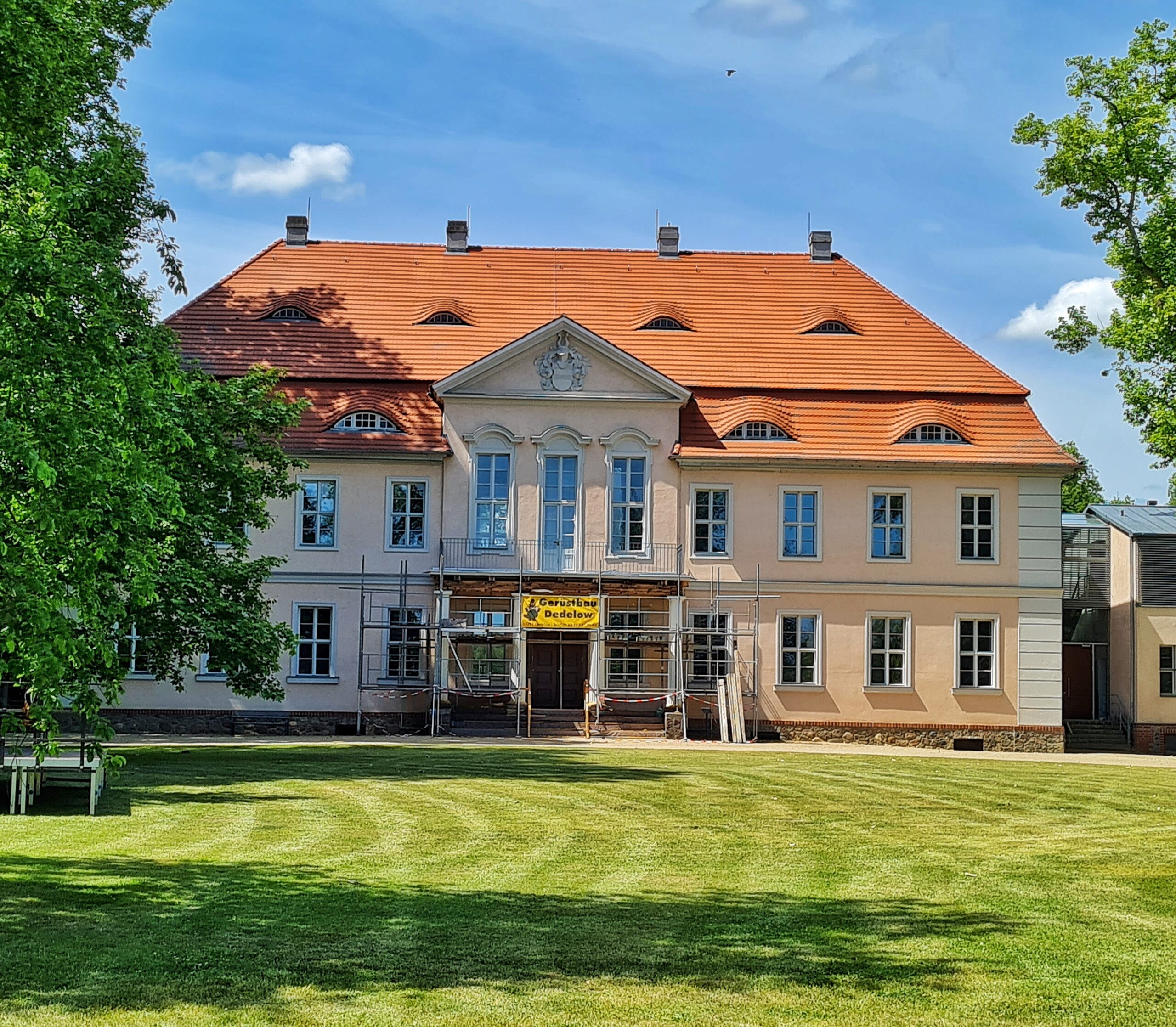 Ein zweigeschossiges helles Gebäude mit vielen hohen Sprossenfenstern und einem hohen roten Dach. Im Eingangsbereich steht ein Baugerüst. Davor eine gepflegte Rasenfläche. Links und rechts ragen Bäume in das Bild.