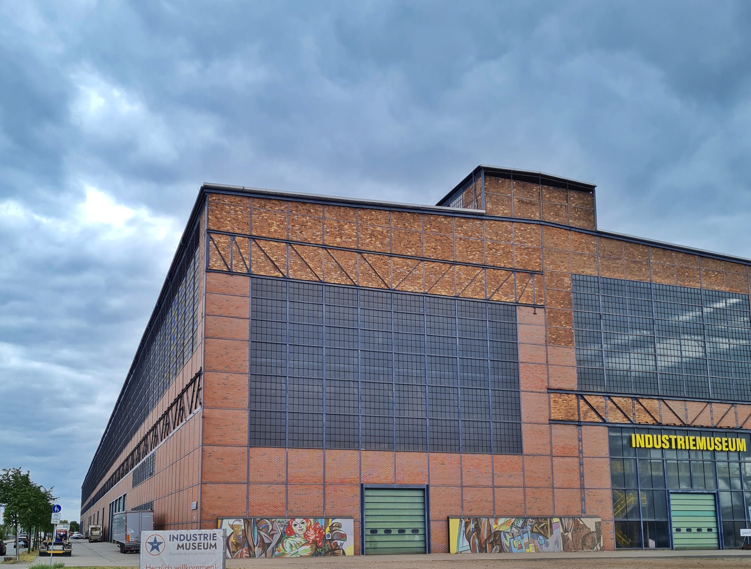 Eine riesige rötliche Fabrikhalle, etwas seitlich fotografiert, so dass man die Vorderseite und die linke Gebäudeseite sieht. Sie hat große Fensterflächen mit Gitter. Vorne links steht ein Schild mit der Aufschrift "Industriemuseum" Die gleiche Aufschrift steht in gelben Buchstaben über dem verglasten Eingangsbereich.