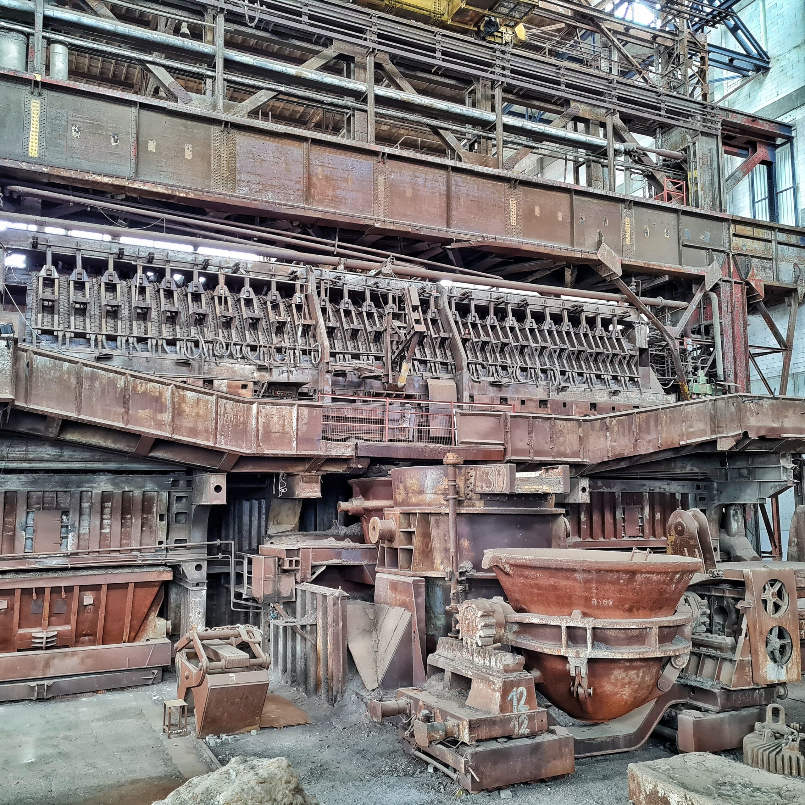 Die andere Seite dses Siemens-Martin-Ofens. Ganz oben ist ein Metallumlauf der begehbar ist. Daruner eine lange Reohe von dicht an dicht liegenden Metallteilen, senkrecht angeordnet. Ganz unten auf dem Boden stehen große Formen, in denen riesige hakbrunde Gefäße befestigt sind.