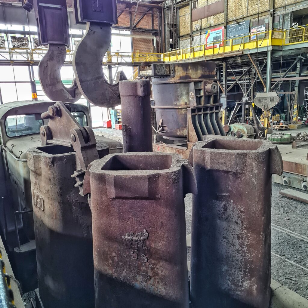 Links eine alte Metalllok, über der zwei sehr große Metallhaken hängen. Im Vordergrund drei hohe, viereckige Metallbehälter.