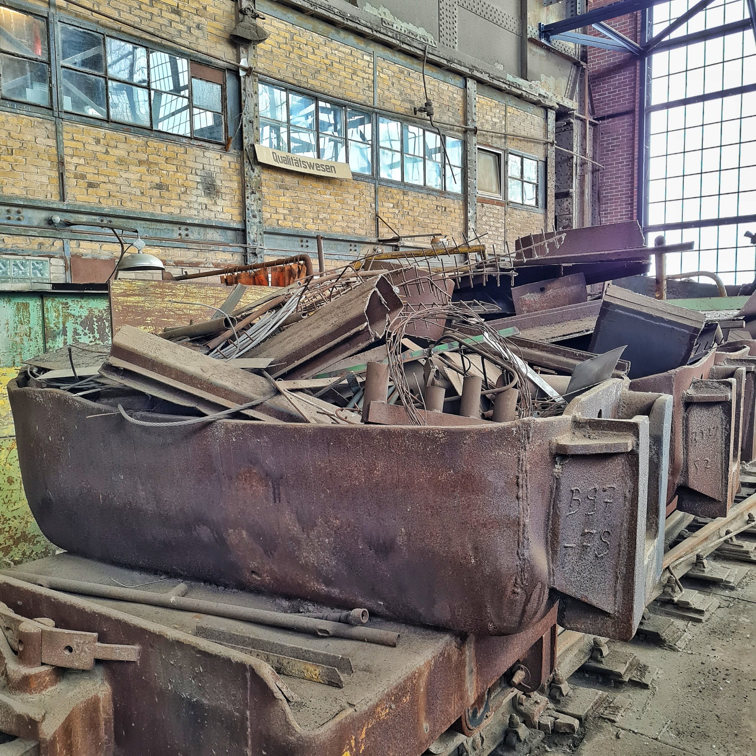 In einer hohen Fabrikhalle stehen mehrere große, längliche Behälter aus Metall schräg auf Gleisen. Sie sind bis über den Rand gefüllt mit alten Metallteilen.