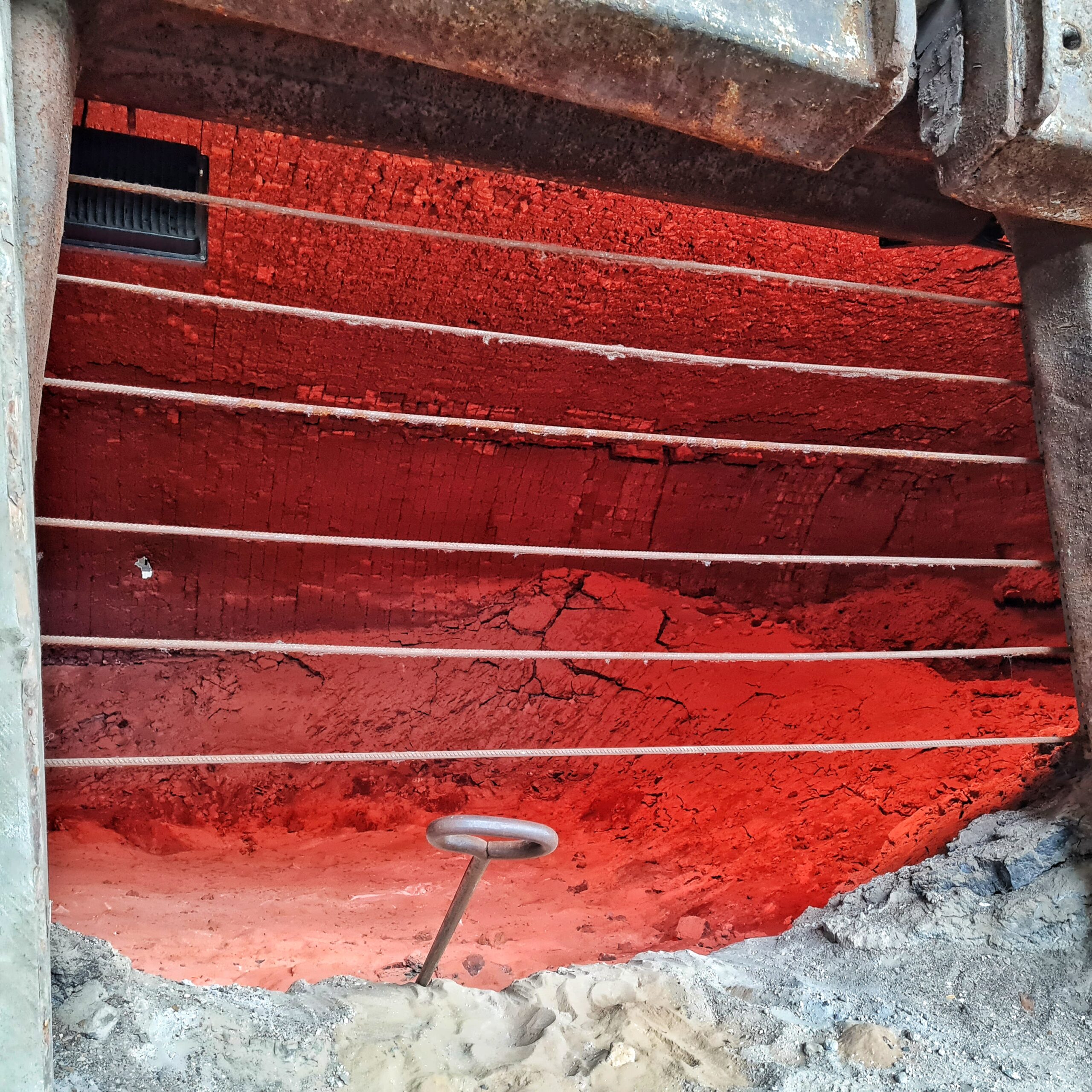 Blick in den Schmelzofen. Darin eine Mulde aus Gestein oder ähnlichem, das Risse hat. Der Ofen ist mit rotem Licht ausgeleuchtet, so dass es aussieht, als ob darin ein Feuer glüht. Vorne sind mehrere parallele Metallstangen waagerecht angebracht. Vorne ragt ein Griff irgendeines Werkzeugs heraus. Es ist eine schmale Stange mit einem Metallkreis am Ende.
