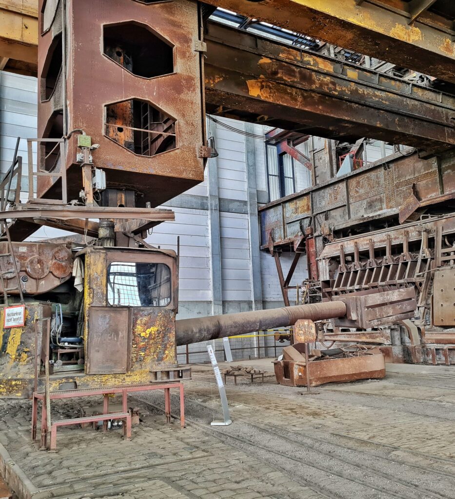 Oben quer durch die Fabrikhalle breite gelbliche Streben. Daran hängt ein längliches Metallteil mit Öffnungen darin. Daran hängt eine Führerkabine, die vorne in Richttung des Siemens-Martin-Ofens ein langes Metallrohr hat, an dessen Spitze ein drehbarer Metallbehälter befestigt ist. Unter der Führerkabine ist ein Metallgestell mit einigen Stufen angebracht, so dass man hineinklettern kann.