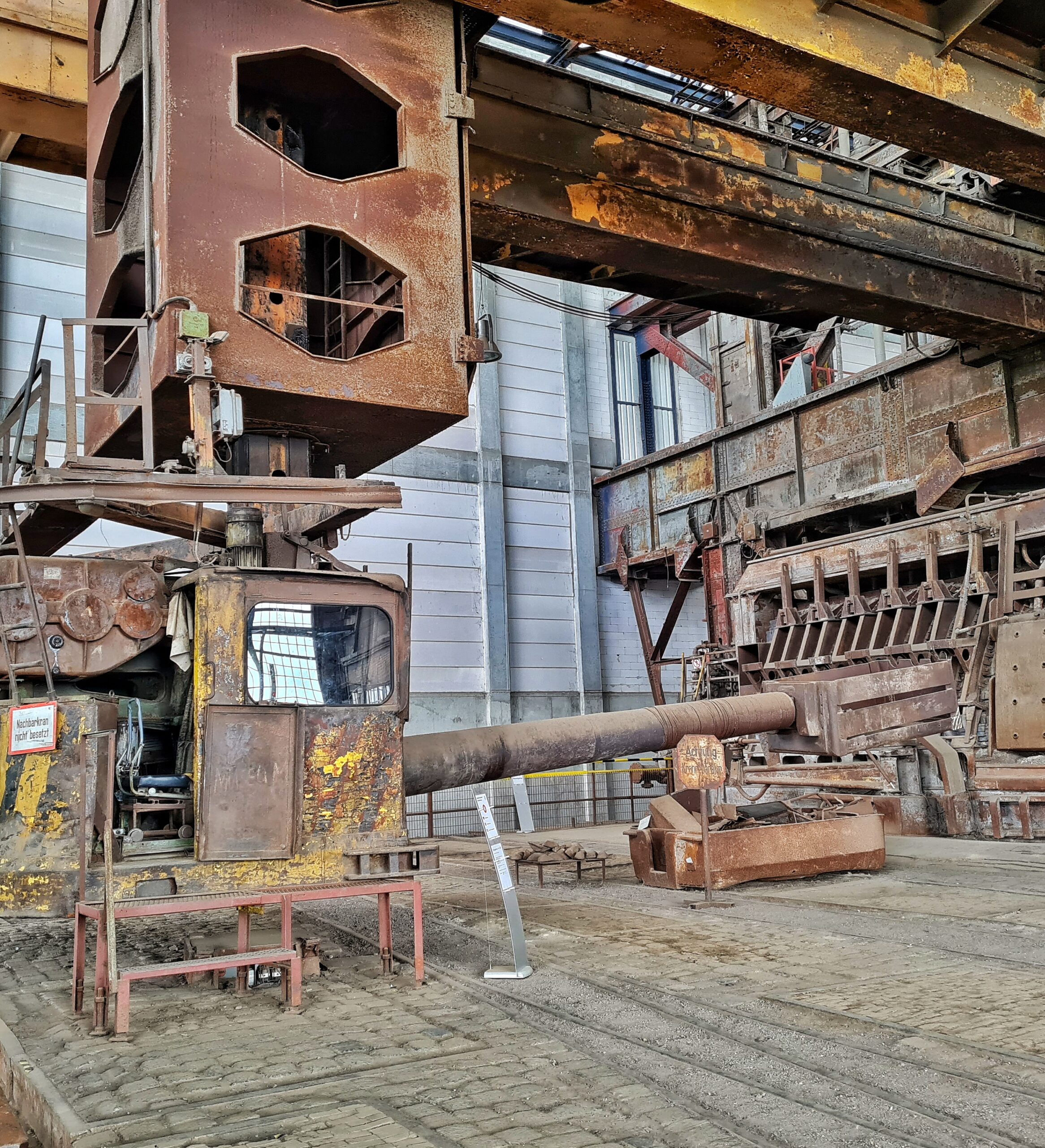 Oben quer durch die Fabrikhalle breite gelbliche Streben. Daran hängt ein längliches Metallteil mit Öffnungen darin. Daran hängt eine Führerkabine, die vorne in Richttung des Siemens-Martin-Ofens ein langes Metallrohr hat, an dessen Spitze ein drehbarer Metallbehälter befestigt ist. Unter der Führerkabine ist ein Metallgestell mit einigen Stufen angebracht, so dass man hineinklettern kann.