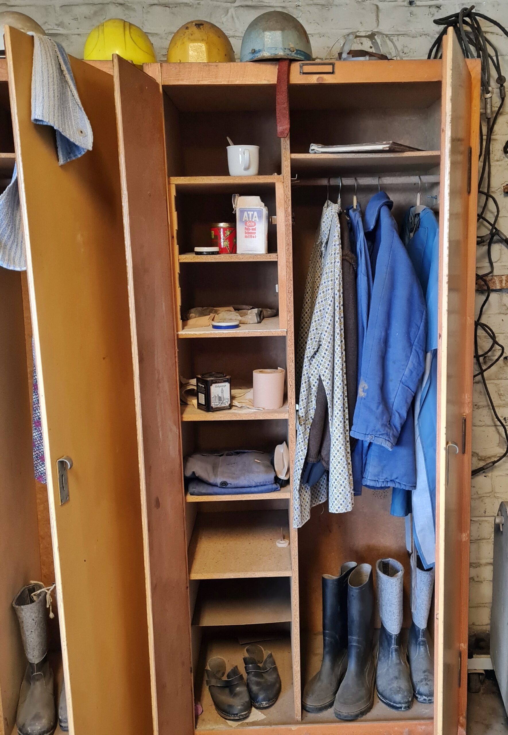 Ein schmaler Schrank mit geöffneten Türen. Obendrauf mehrere Arbeitshelme. Rechts hängen vier Hemden oder Jacken. Darunter stehen zwei Paar hohe Stiefel. In den Fächern auf der linken Seite liegen weitere Hemden, und es stehen dort Tassen, Ata, eine Cremdeose, Klopapier und Schuhe. Über der linken Tür hängt ein Handtuch.
