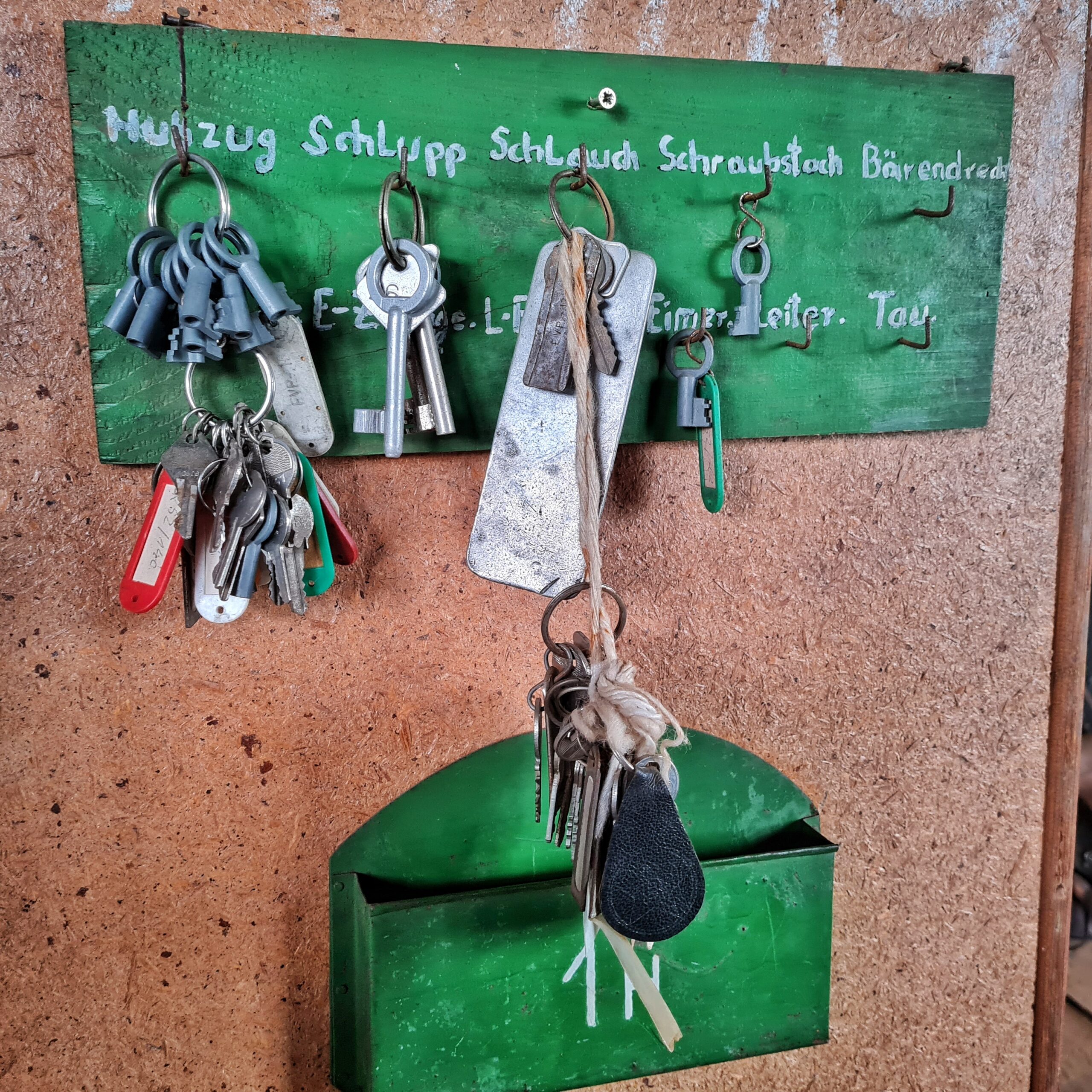 Ein grünes Schlüsselbrett aus Metall hängt an einer Wand, die aussieht wie eine dunkle Spanplatte. An mehreren Haken hängen eimzelne Schlüssel oder Schlüsselbunde. Die Haken sind in weißer Schrift beschriftet, wobei dies teilweise durch die Schlüssel verdeckt ist. Sichtbar sind die Aufschriften: Schlupp, Schlauch, Schraubstock, Bärendreck, Eimer Leiter, Tau. Darunter hängt ein schmales grünes Metallgefäß, möglicherweise für Post.