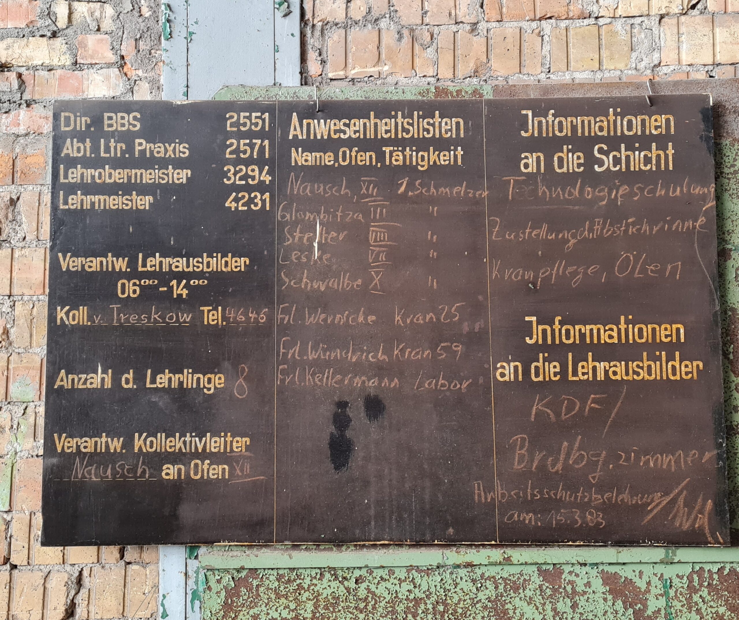 An einer gelblichen Mauer ist eine dunkle Tafel befestigt. Auf der linken Seite stehen oben die vierstelligen Telefonnummern des Dir. BBS, des Abt. Ltr. Praxis, des Lehrobermeisters und des Lehrmeisters. Darunter steht Verantwortlicher Lehrausbilder 6 - 14 und darunter ist mit Kreide der Name v. Treskow und eine vierstellige Nummer geschrieben. Darunter steht Anzahl der Lehrlinge, dahinter mit Kreide 8. Darunter steht Verantw. Kollektivleiter und darunter mit Kreide Nausch an Ofen XII. In der Mitte steht: Anwesenheitslisten Name Ofen, Tätigkt. Darunter mit Kreide mehrere Nachnamen, die Nummer des jeweiligen Ofens und 1. Schmelzer. Darunter mit Kreide: Frl. Wernicke Kran 25, Frl. Wündrich Krank 59, Frl. Kellermann Labor. Auf der rechten Seite steht: Informationen an die Schicht, darunger mit Kreide: Technologieschulung, Zustellung d. Abstichrinne, Kranpflege, Ölen. Darunter steht: Informationen an die Lehrausbilder, darunter mit Kreide: KDF/Brdbg.zimmer, Arbeitsschutzbelehrung am 15.3.83